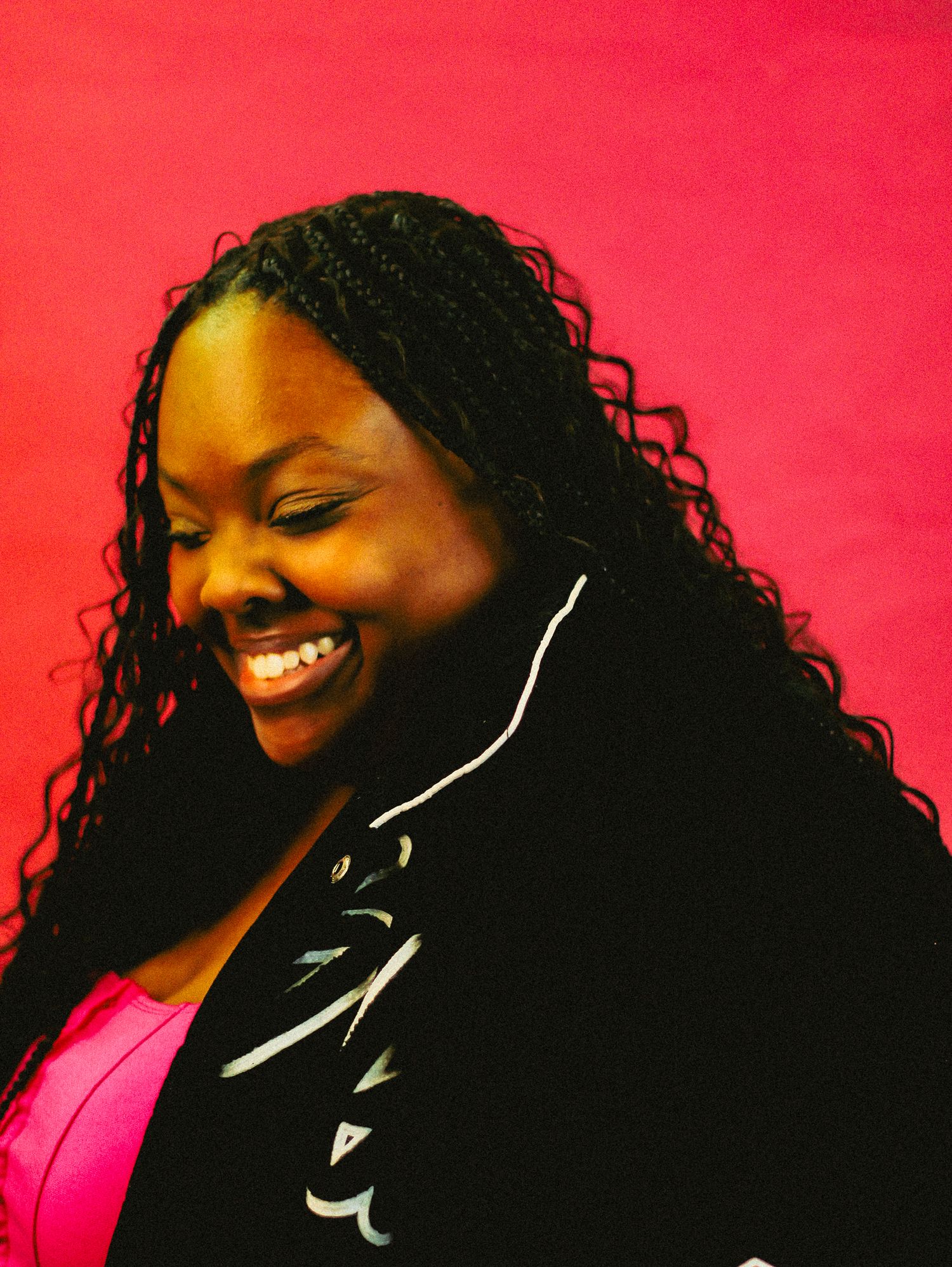 Woman with long braided hair laughs joyfully wearing a black jacket over pink top against a bright pink background.