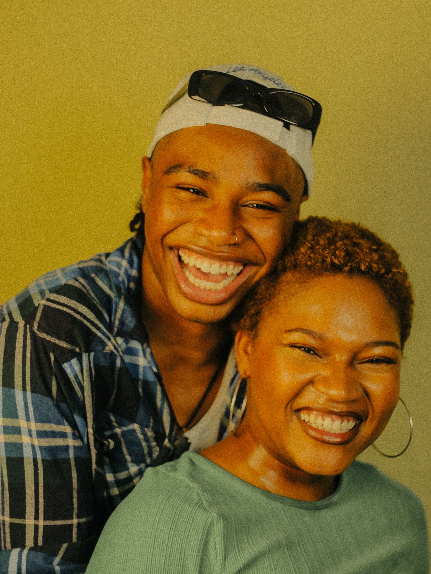 Two smiling young people posing together against yellow background, one in blue plaid shirt and white cap.
