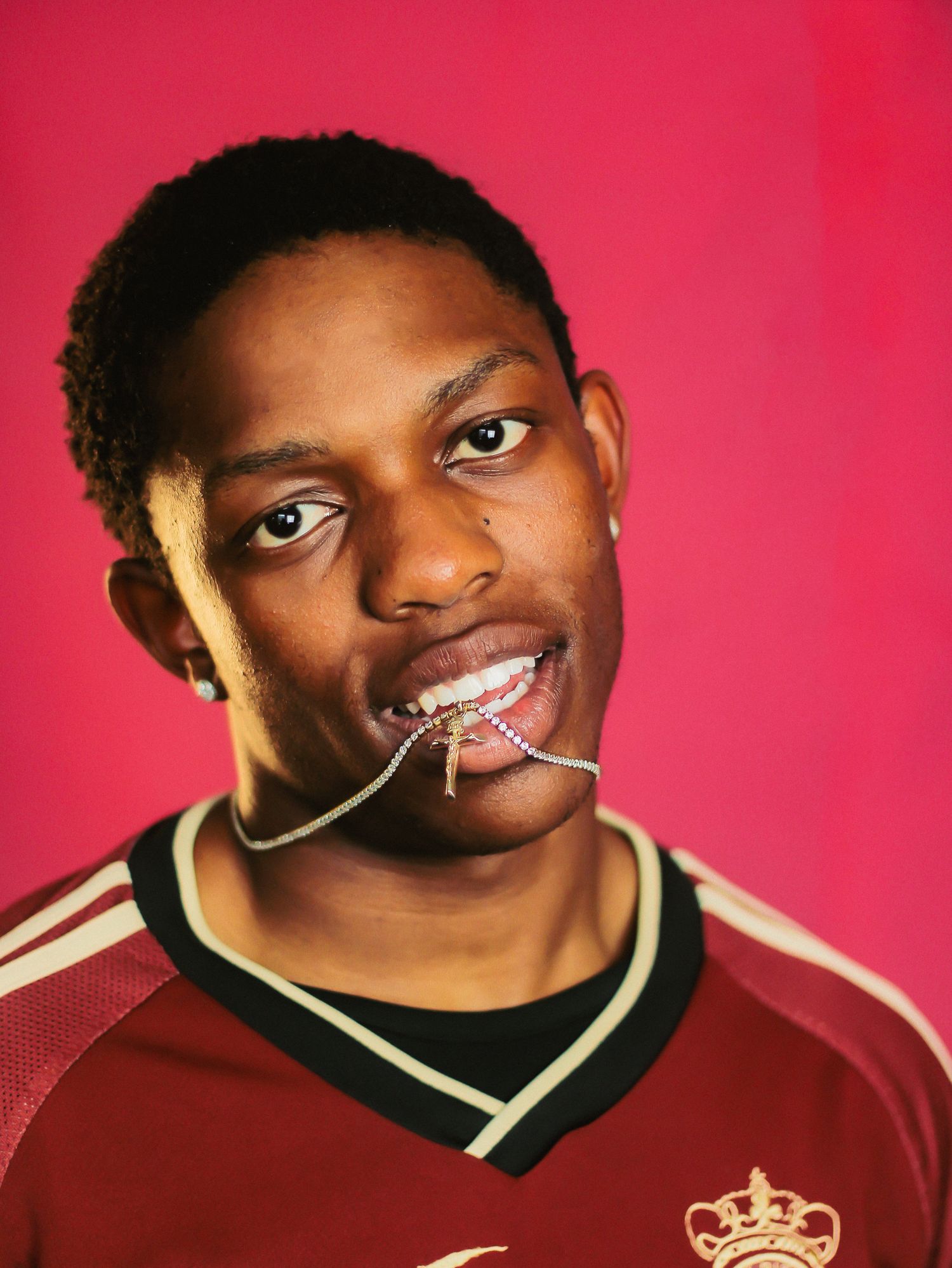 Young Black man in maroon jersey holds chain in mouth against bright pink background.