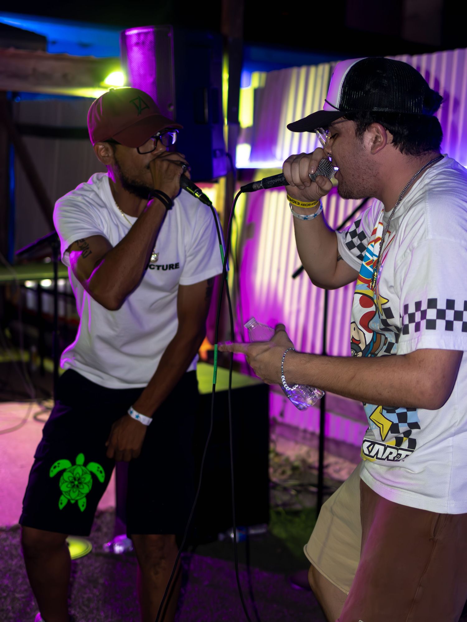 Performers in casual attire singing on stage with vibrant neon lighting effects.
