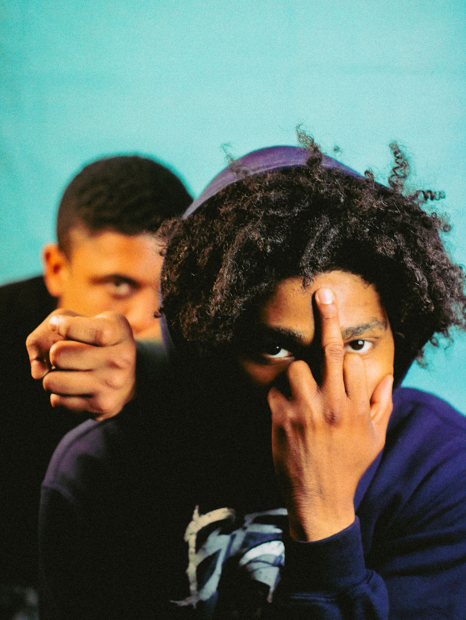 Two young men posing together against turquoise background, one covering face with hand.