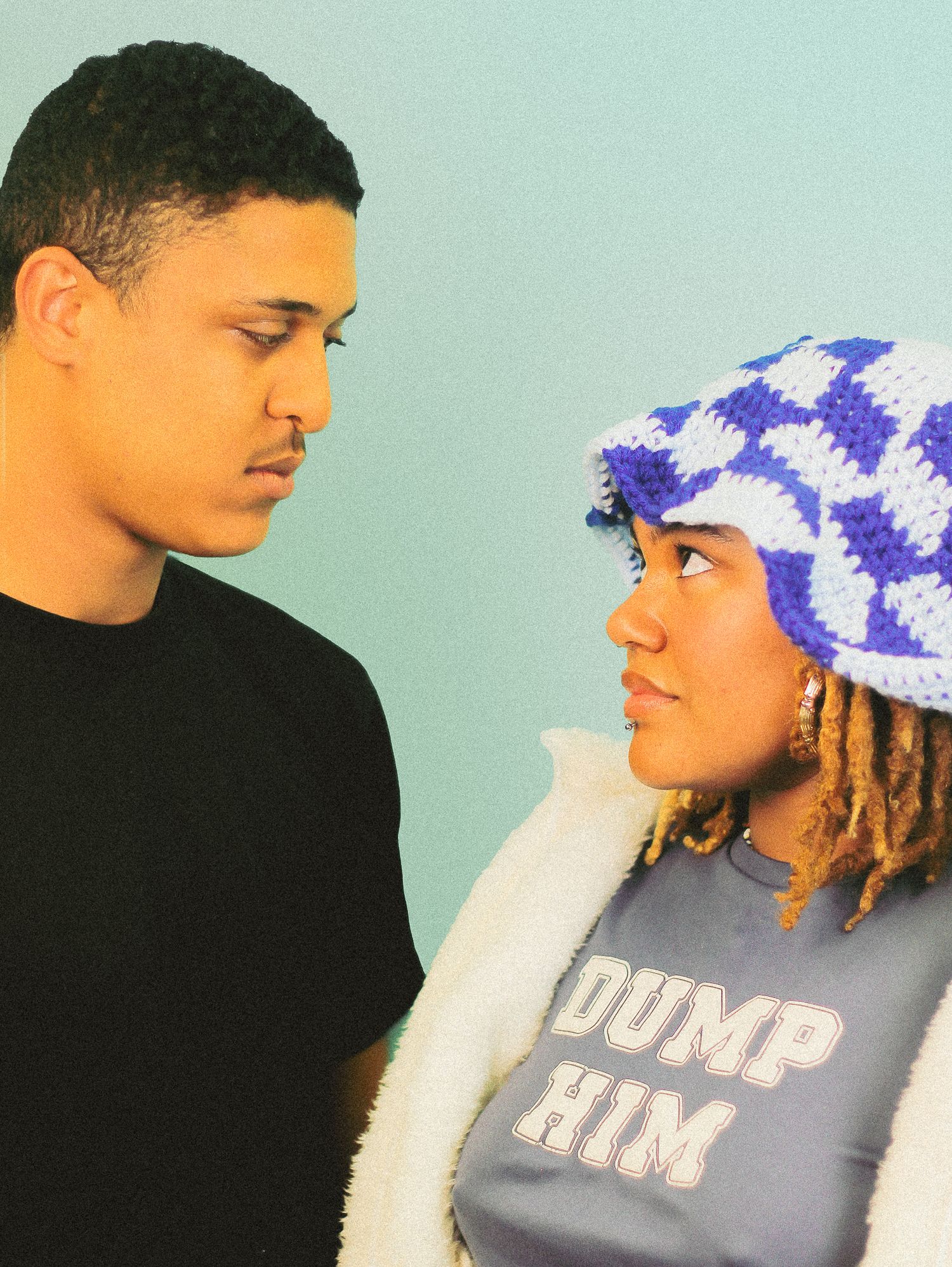 Young man in black shirt and woman wearing blue checkered bucket hat exchange glances on teal background.