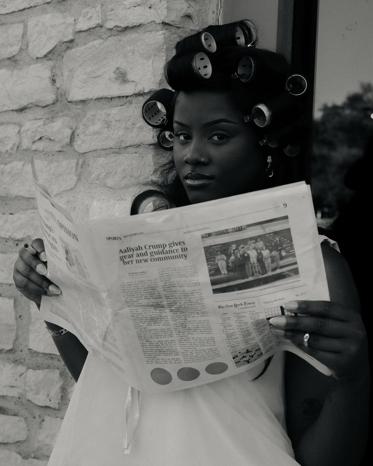 A person with hair rollers reads a newspaper against a white stone wall.