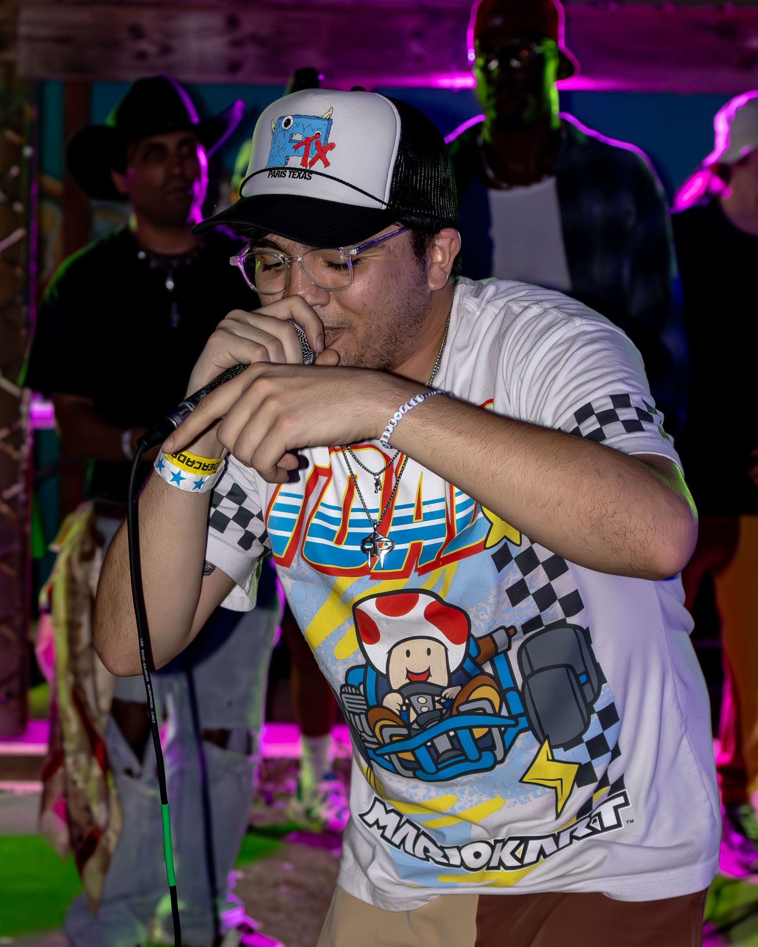 Performer in Mario Kart t-shirt and baseball cap singing into microphone at nighttime event with purple lighting.