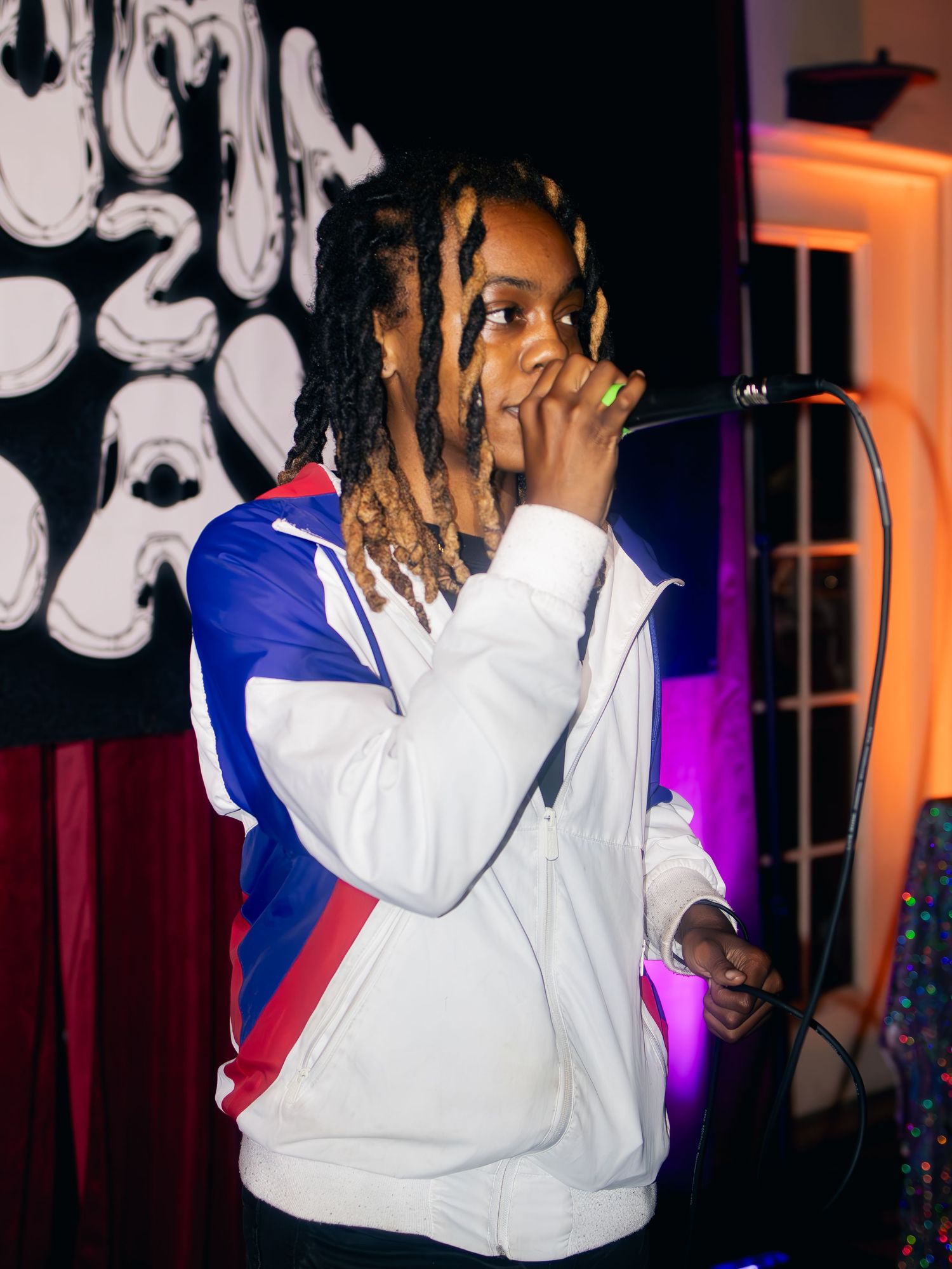 Performer in white and blue track jacket singing on stage with artistic backdrop.