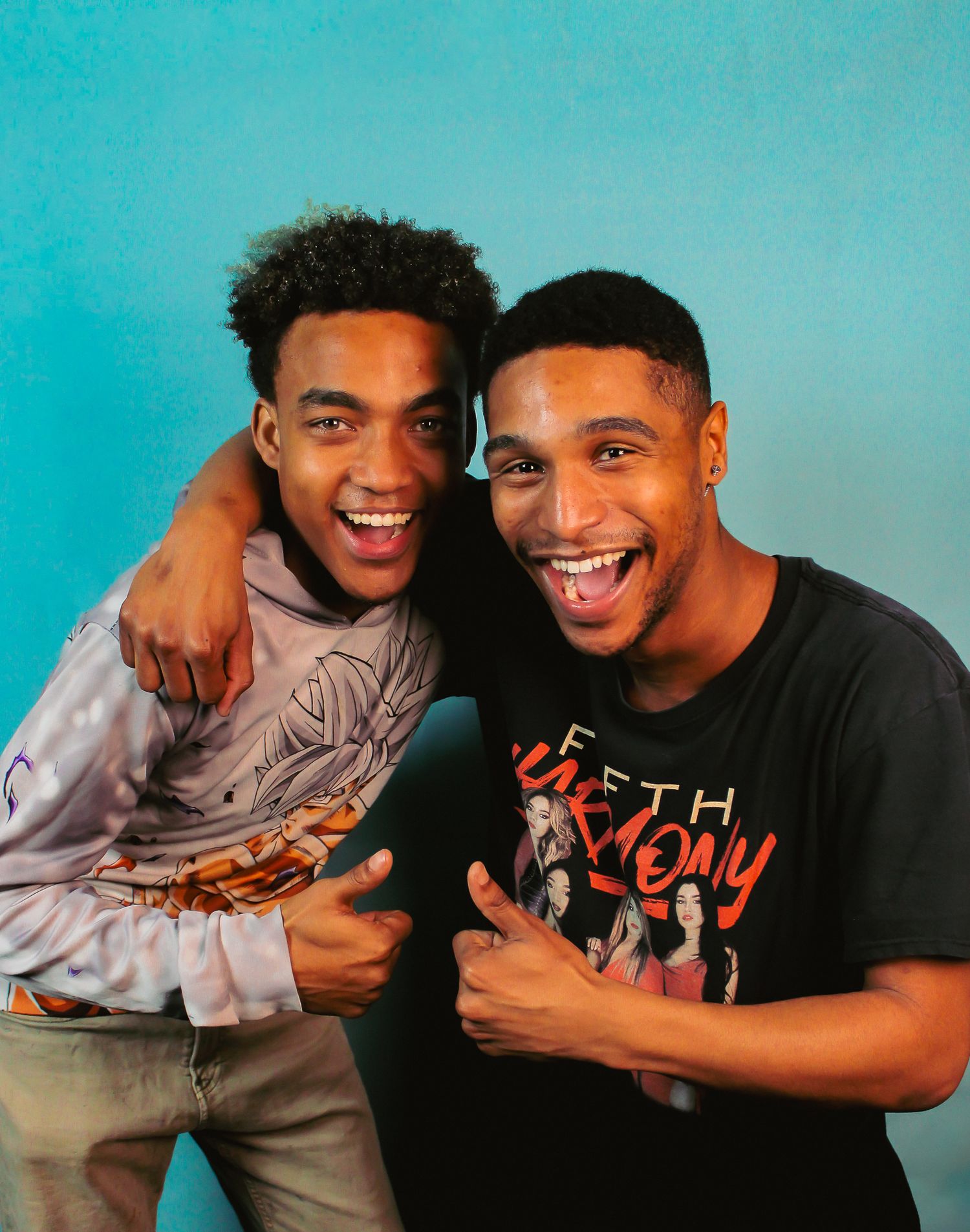 Two friends laughing and embracing while wearing casual clothing against a turquoise background.