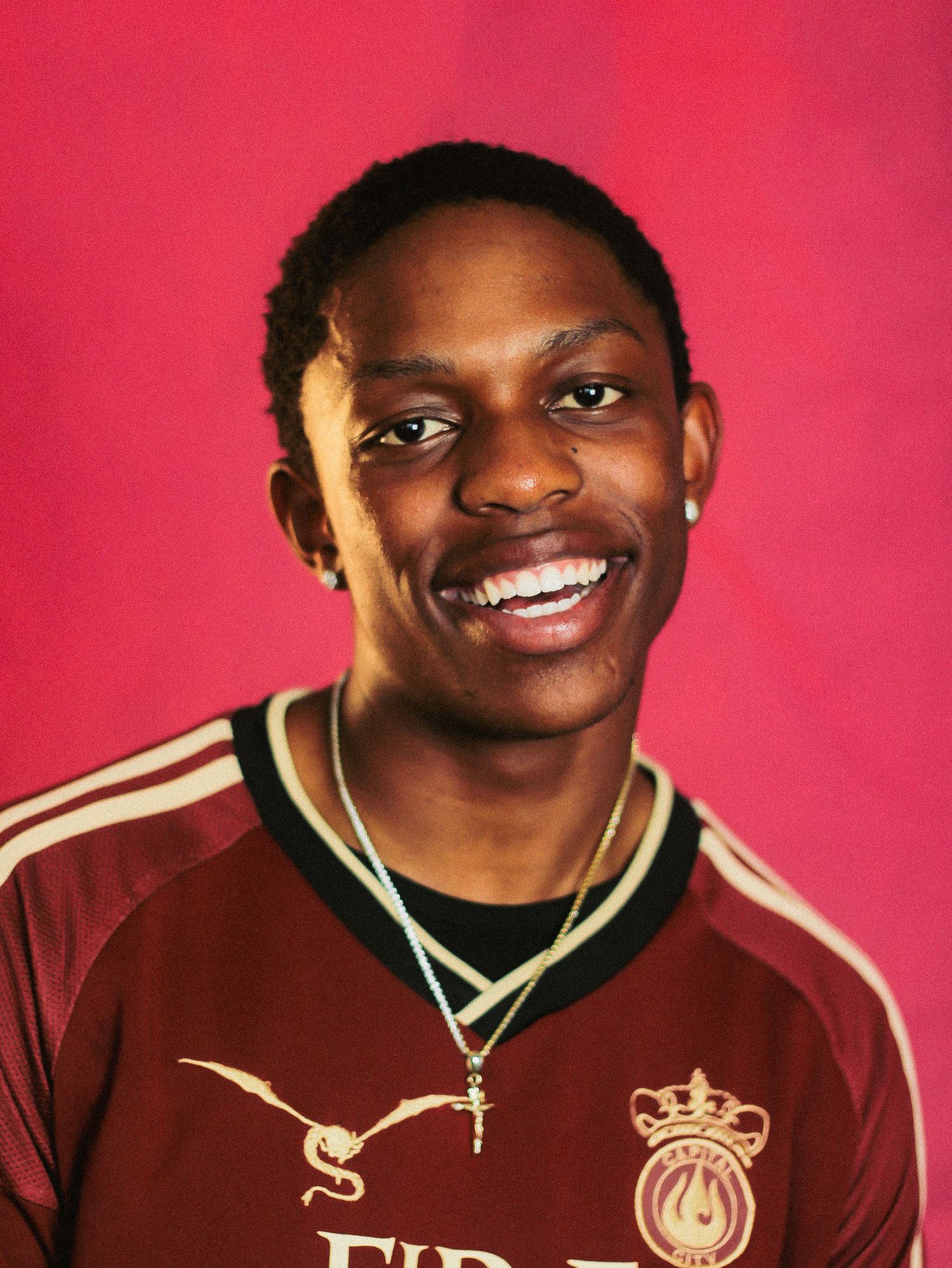 Smiling young Black man in maroon soccer jersey with gold cross necklace on pink background.