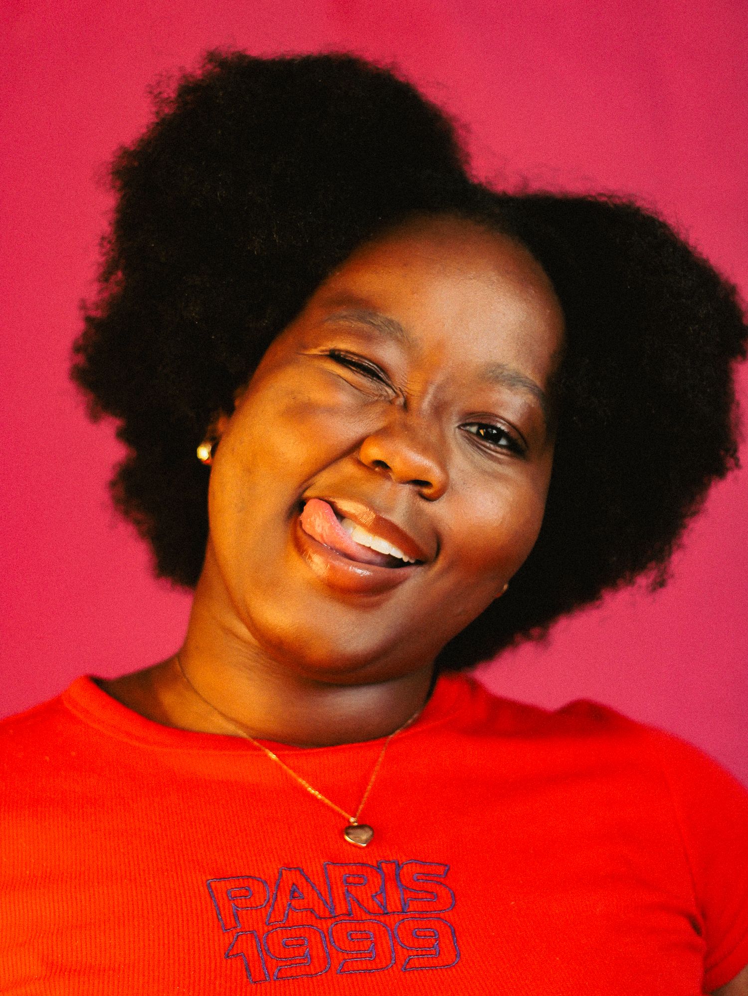 Laughing Black woman with voluminous natural afro hair in a red Paris 1999 shirt on a pink backdrop.