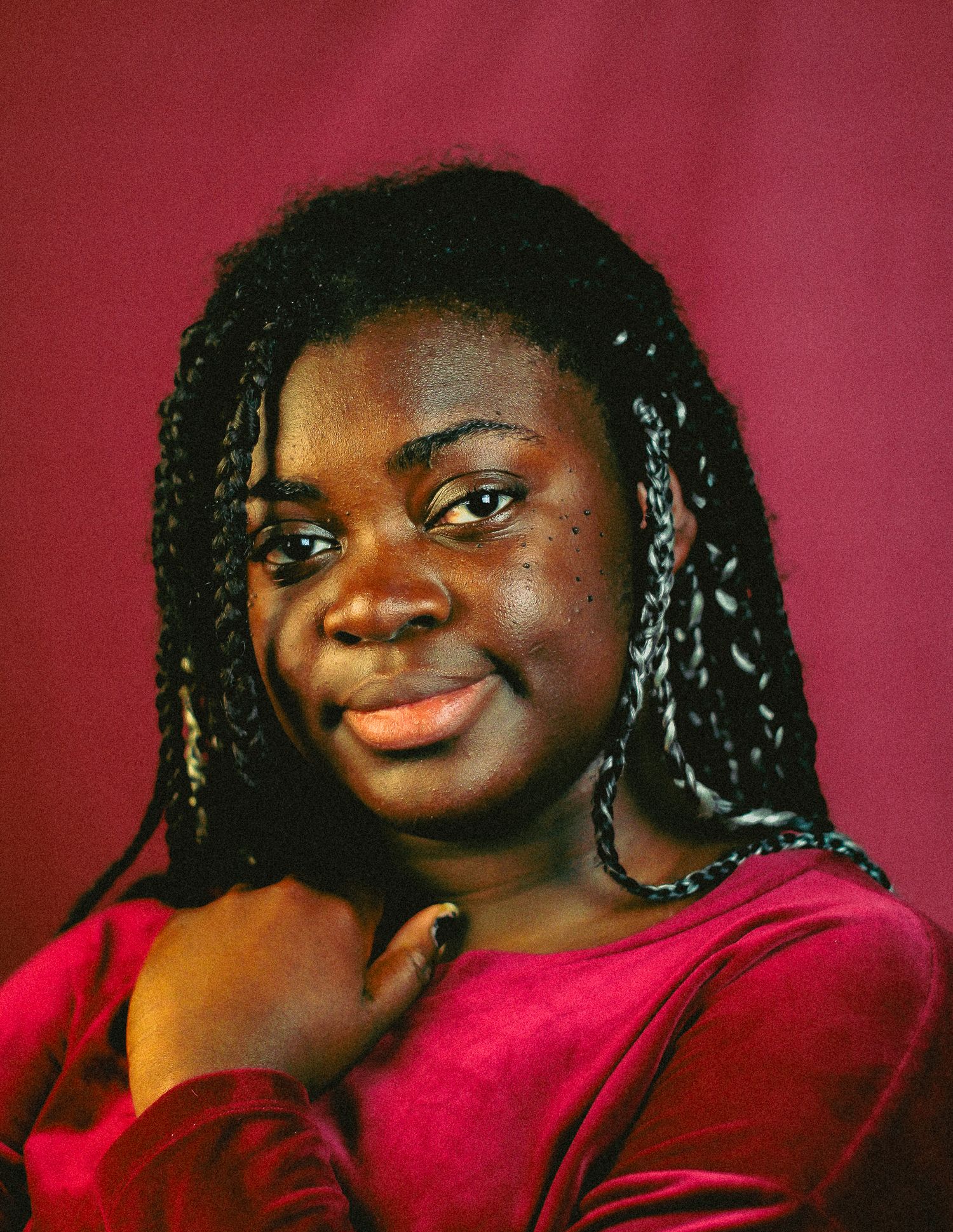 Young woman with braids rests chin on hand, smiling softly at camera against a rich red studio background.