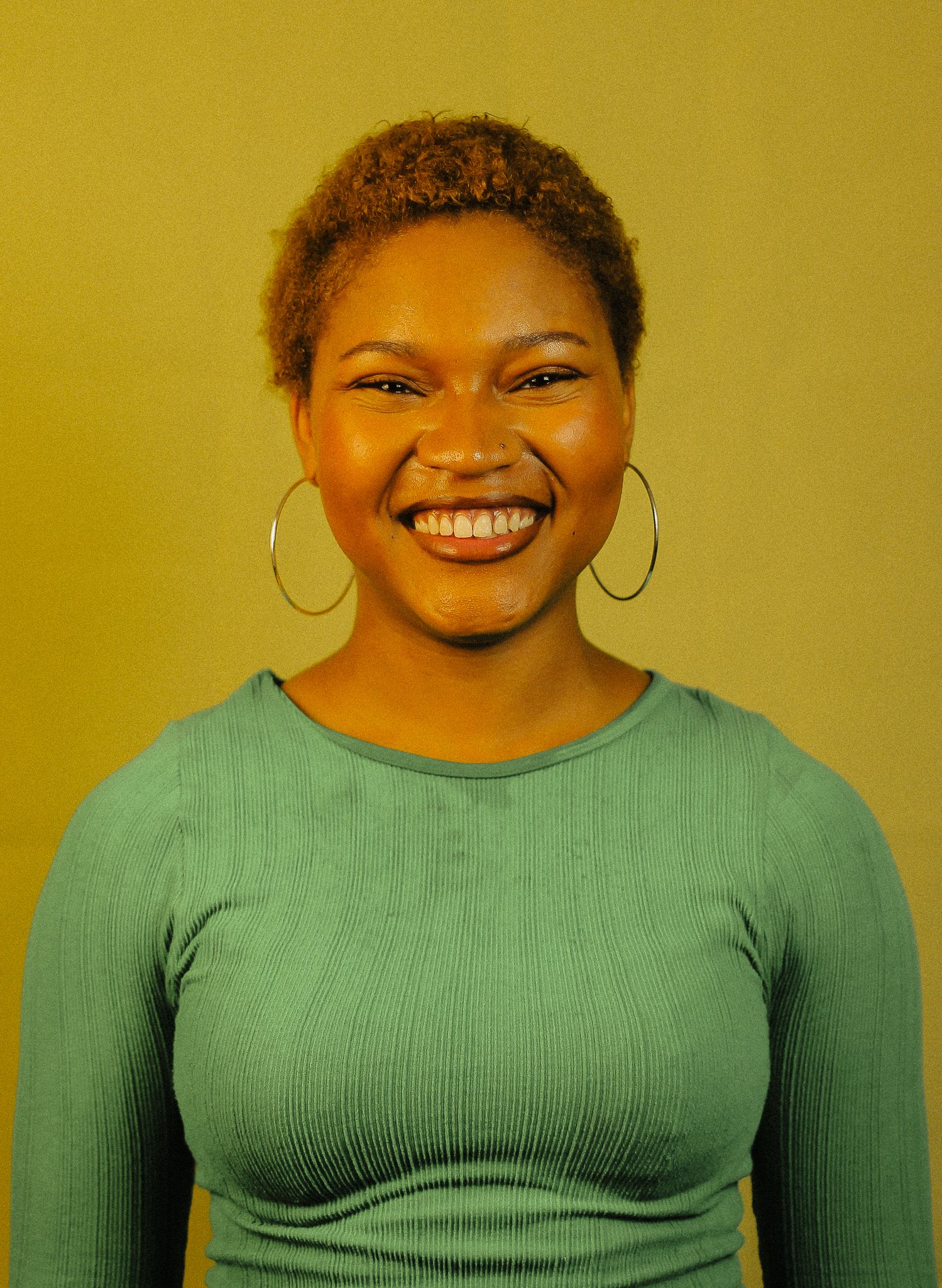 Young woman with short natural hair smiling broadly, wearing teal ribbed long-sleeve top and gold hoop earrings.