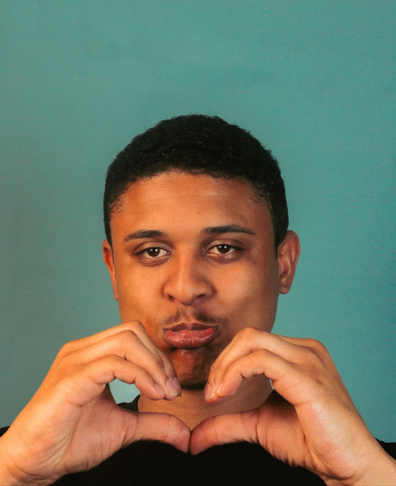 Young man making a heart shape with his hands near his face on teal background.
