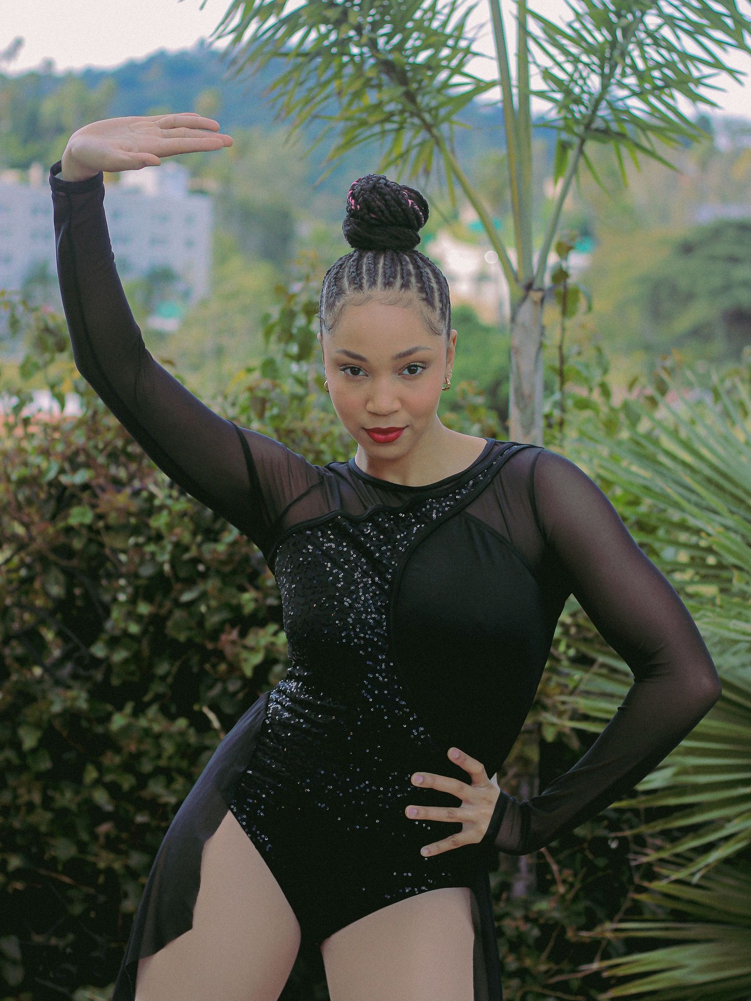 A dancer in a black sequined leotard with sheer sleeves strikes a graceful pose against a tropical garden backdrop.