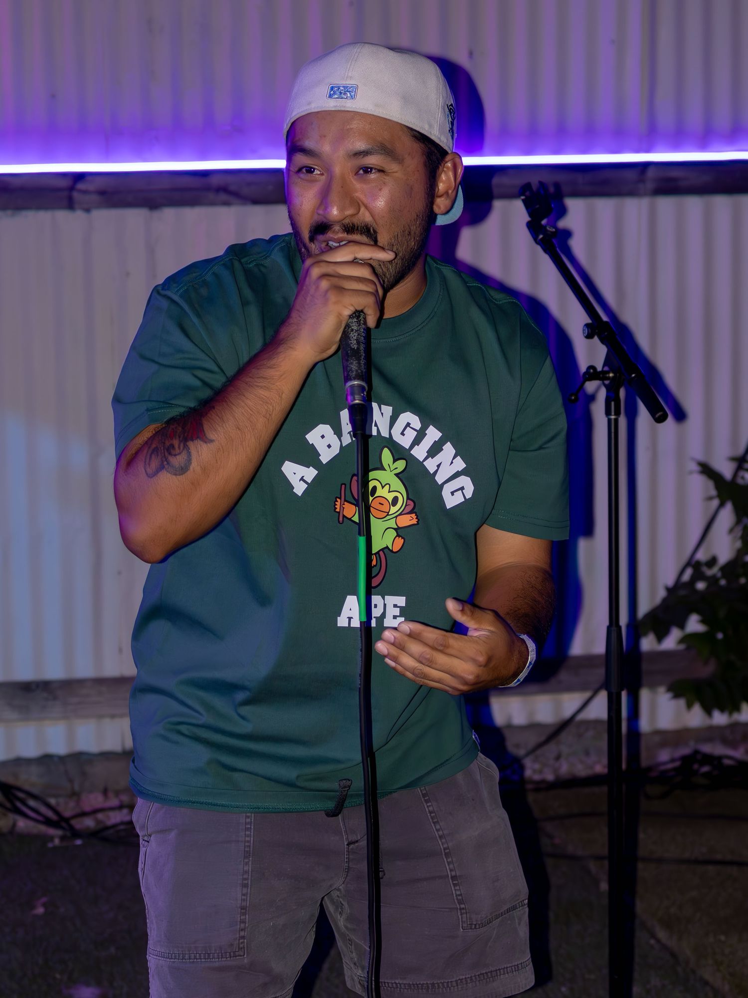 Performer in BAPE green t-shirt and white cap holds microphone during live show.