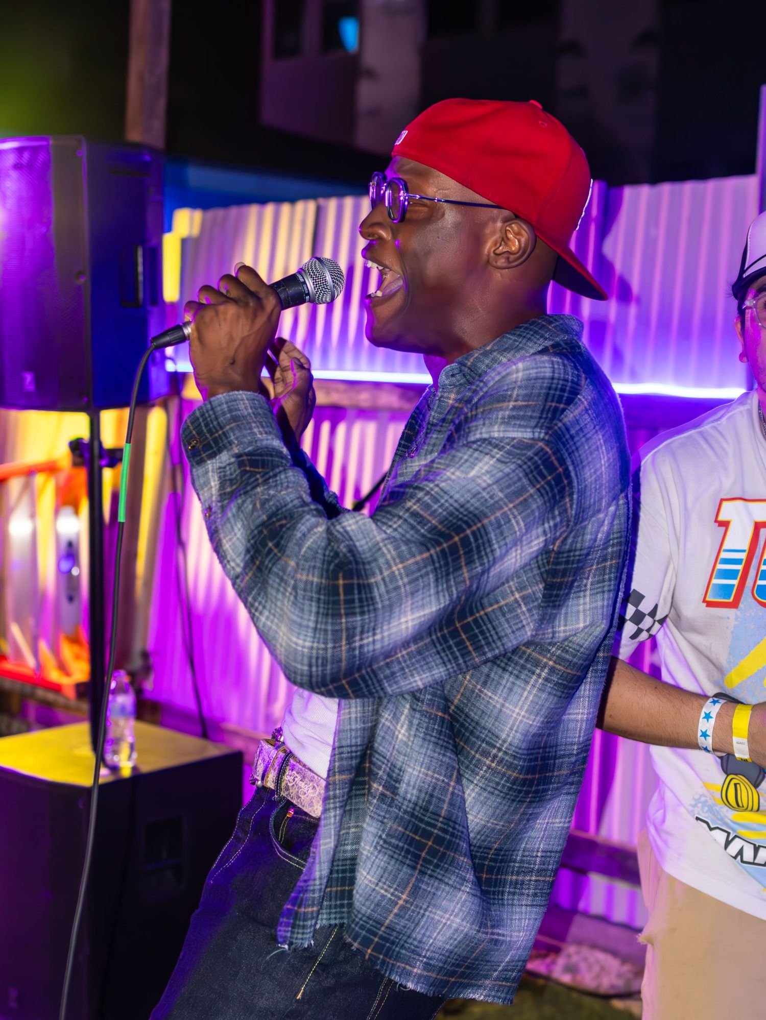 Concert performer in plaid jacket and red cap singing at venue with purple lighting effects.