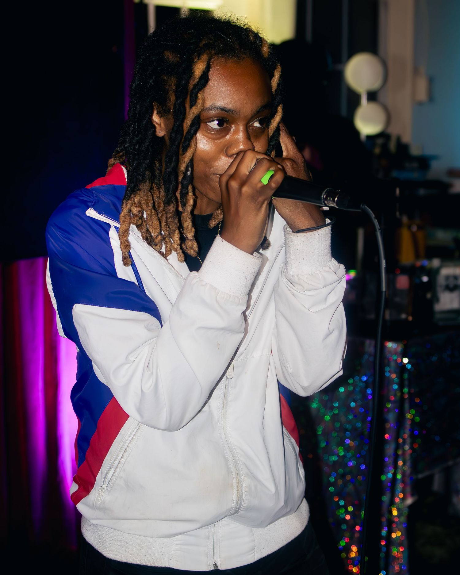Artist in white athletic jacket performing on stage with colorful party lights visible.