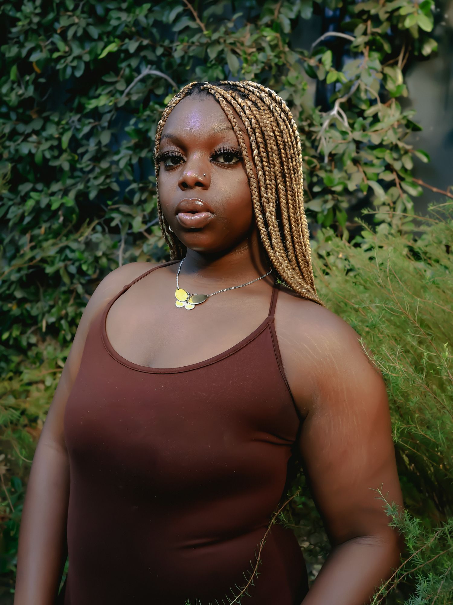 Person in brown tank top poses outdoors against lush green foliage background.