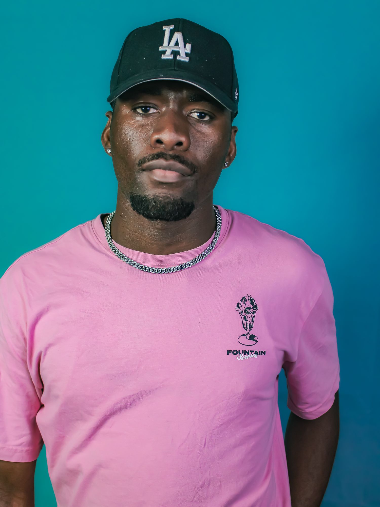 A man in a pink Fountain t-shirt and black LA cap poses against a teal background.
