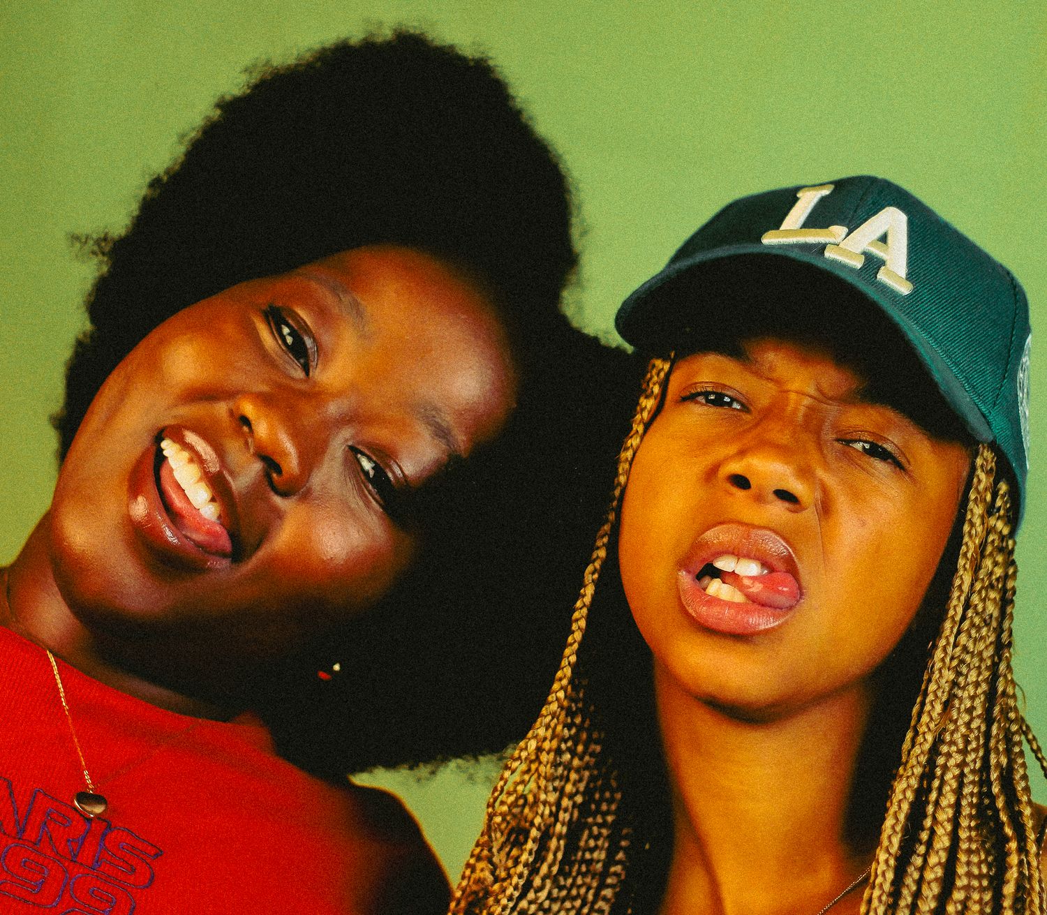 Two young Black women laughing and making silly faces together, one with afro, one wearing green LA cap.