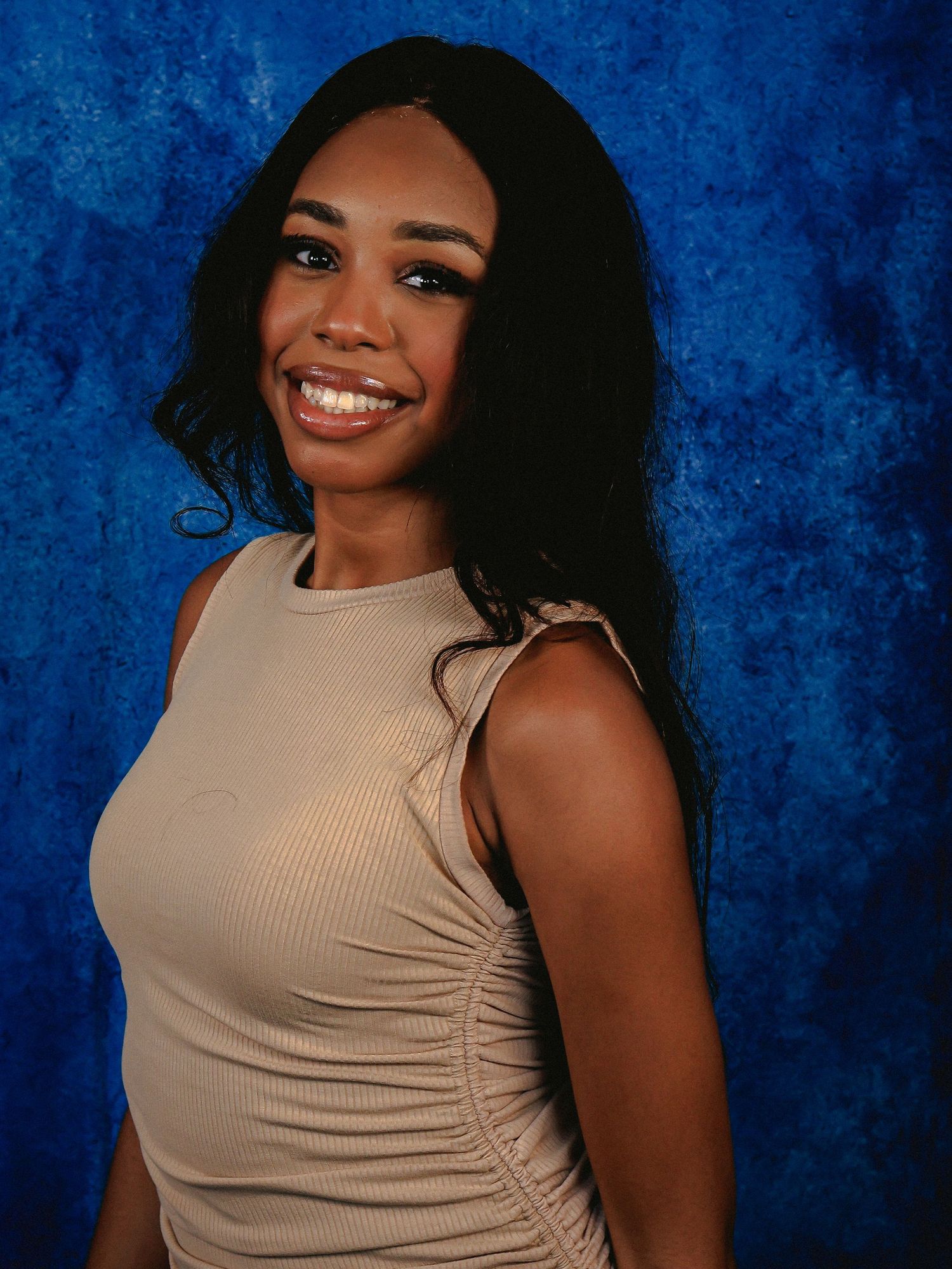 Woman in beige sleeveless top poses confidently against blue background.