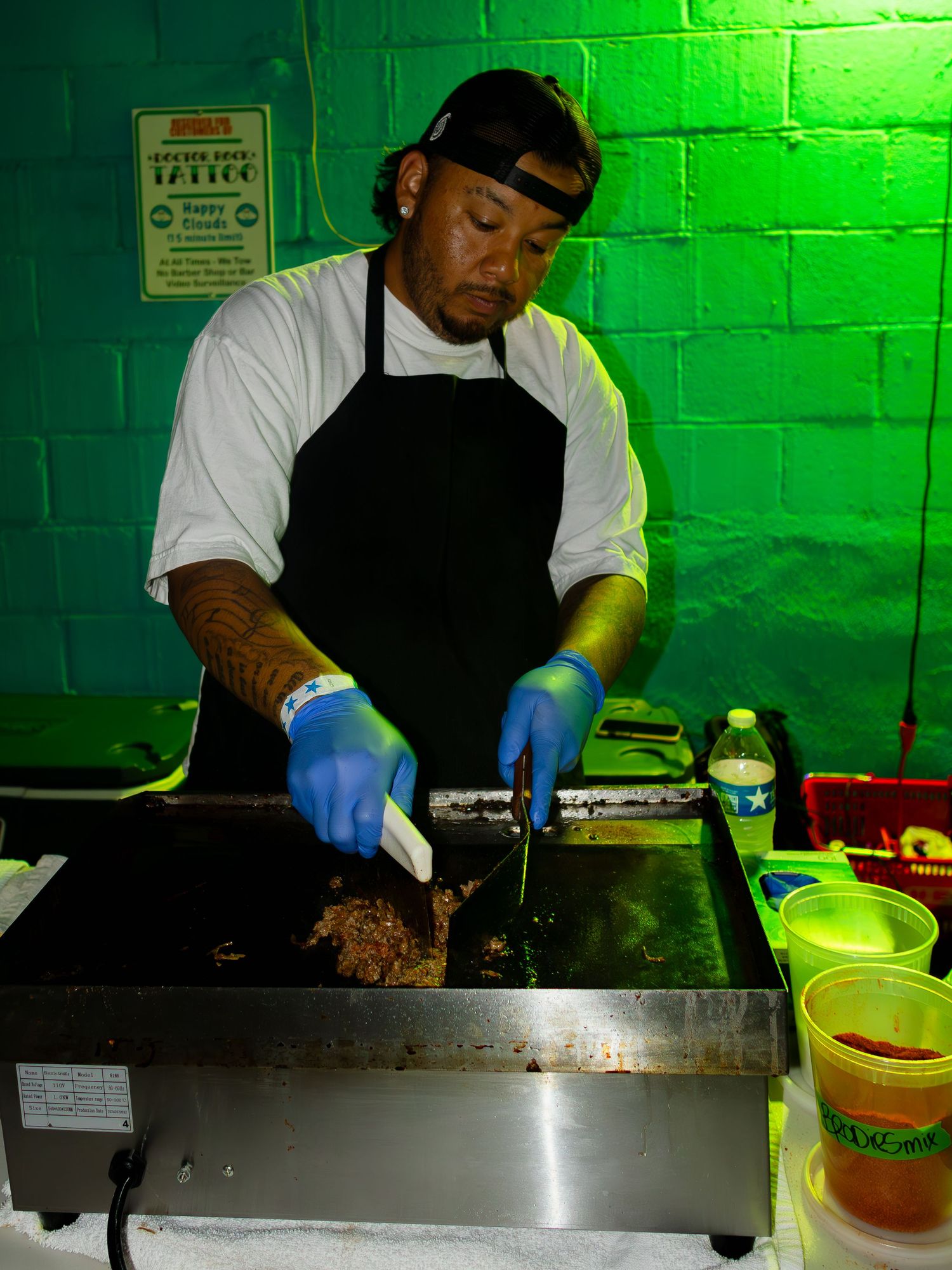 Chef in black apron cooking on a large stainless steel griddle surface with green-lit walls.