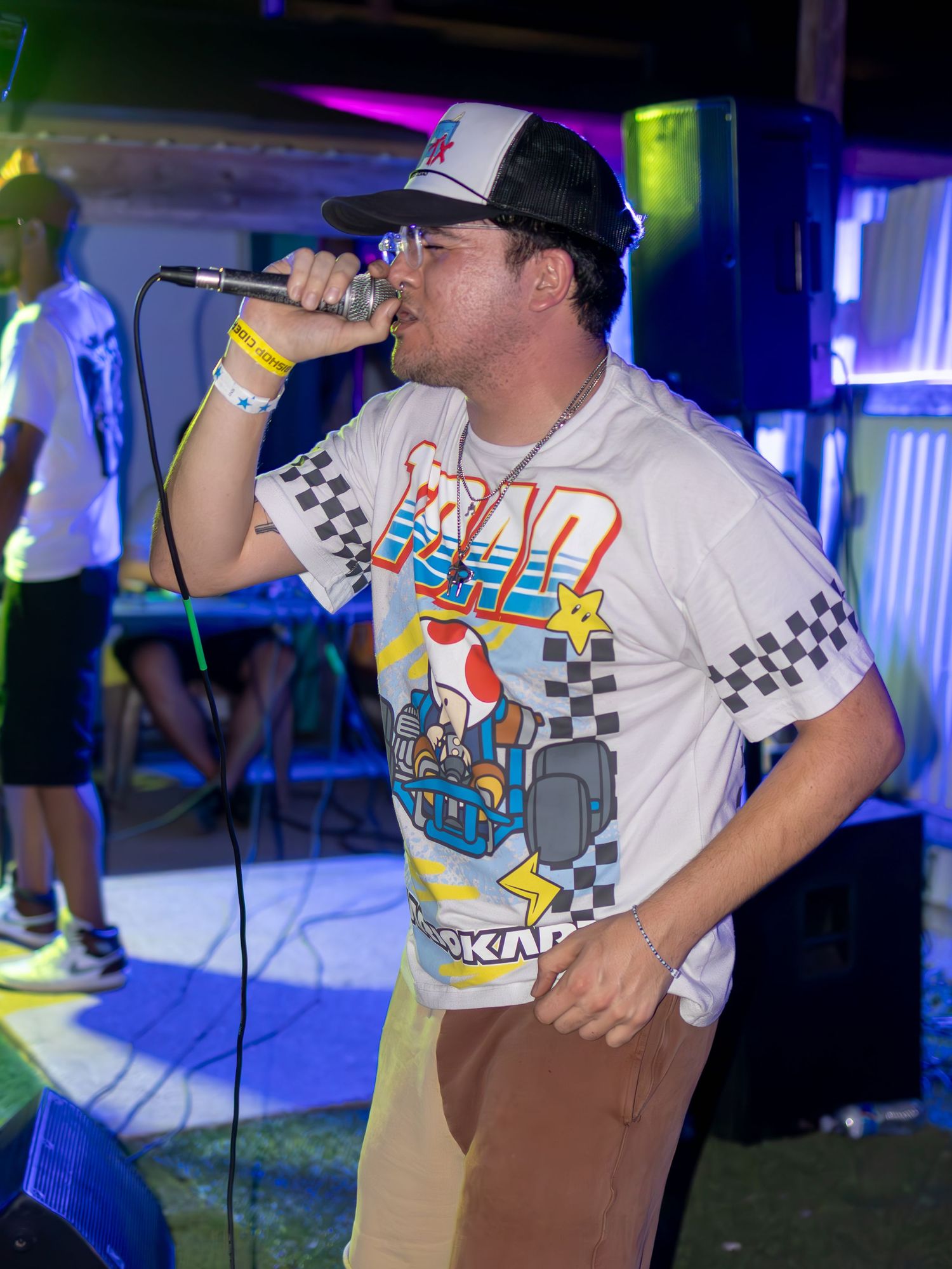 Performers in white t-shirts with checkered patterns sing on stage under colorful nightclub lighting.