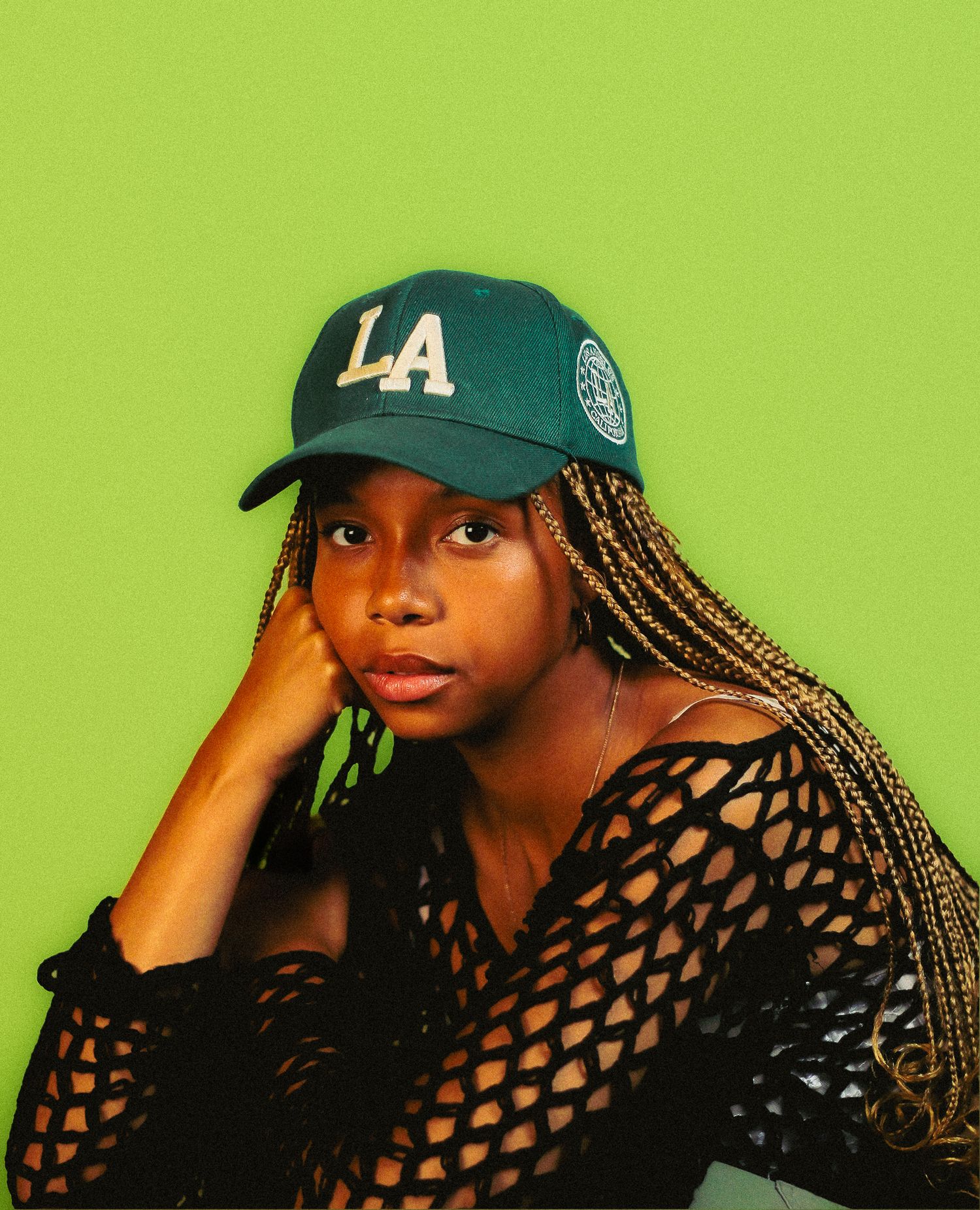 Young woman in LA cap and mesh top leaning on hand against bright green background.