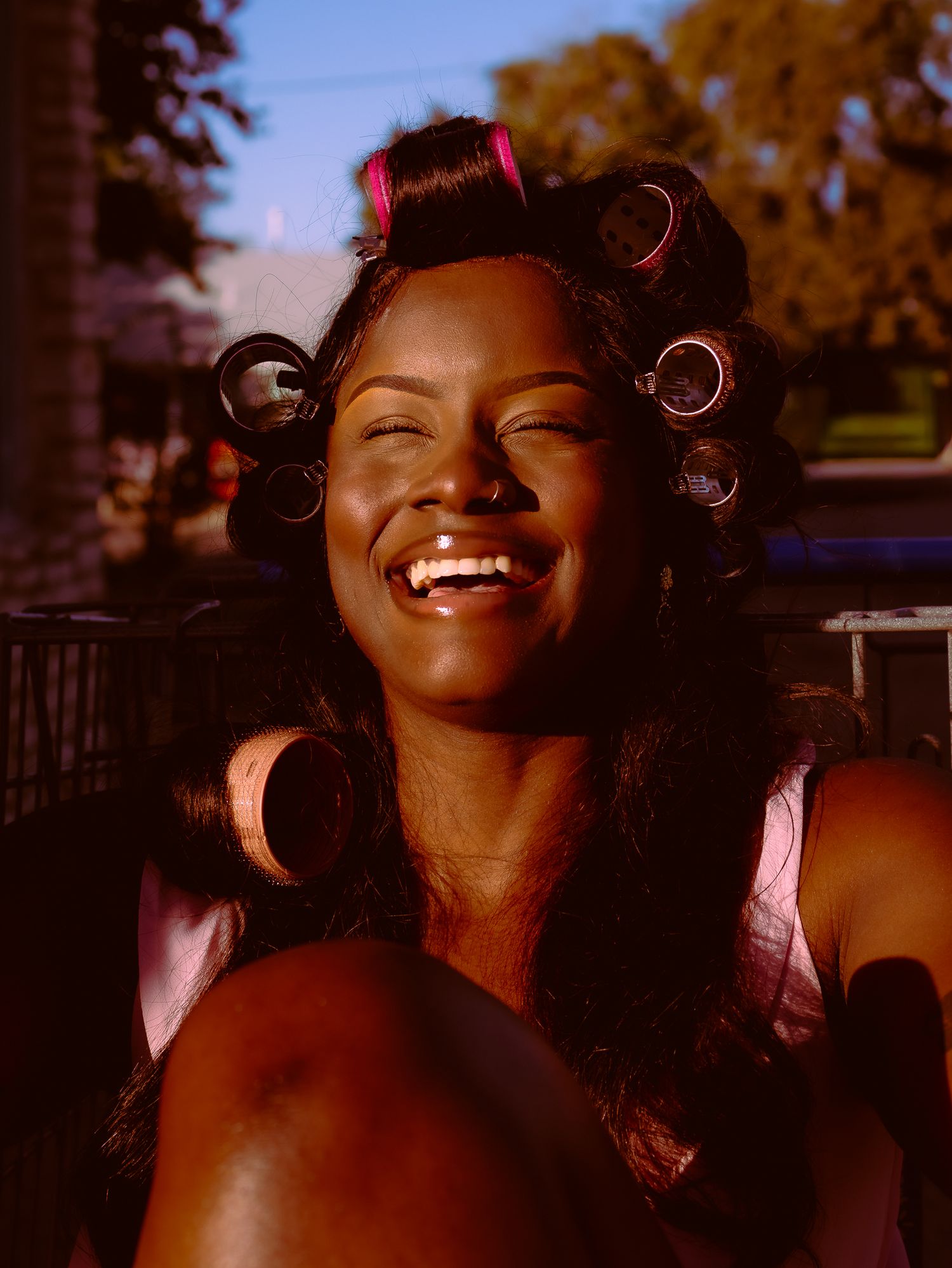 Woman with hair rollers laughs joyfully in warm sunset lighting.