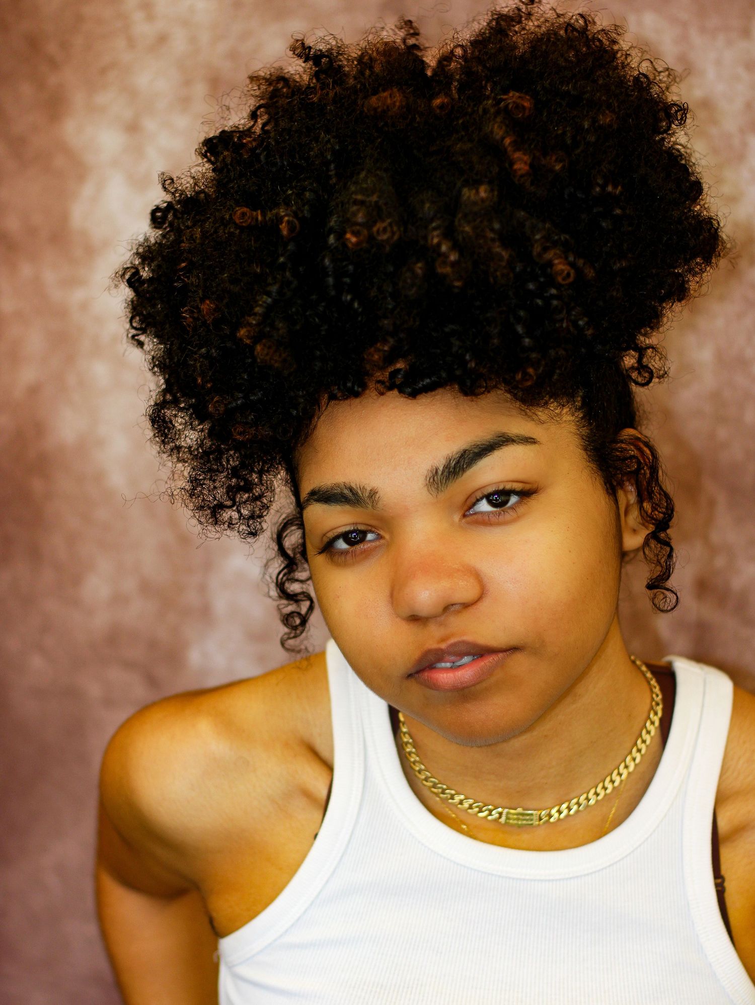 Portrait with natural curly hair and white tank top against brown backdrop.