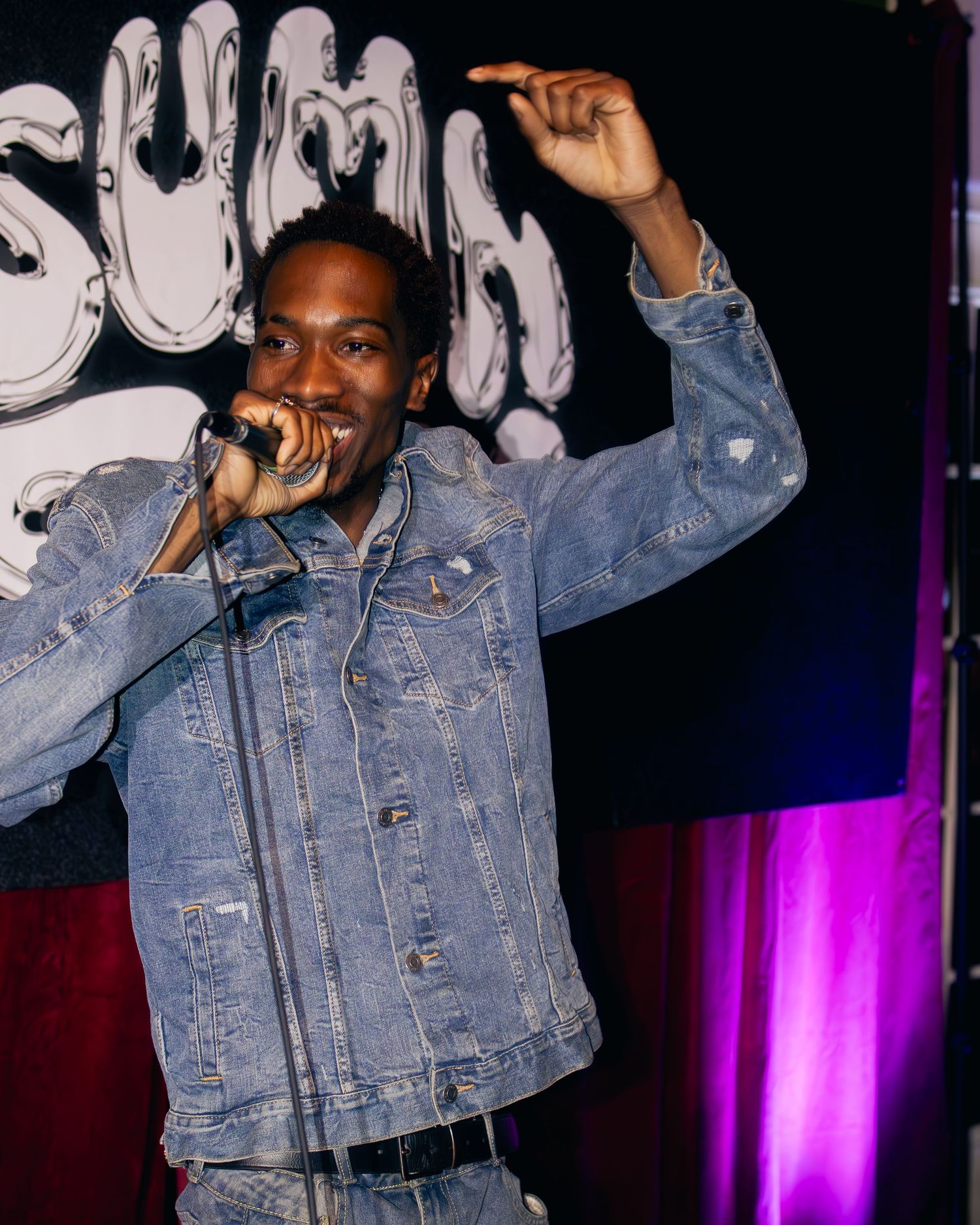 Performer in denim jacket enthusiastically singing during a live performance with purple stage lighting.