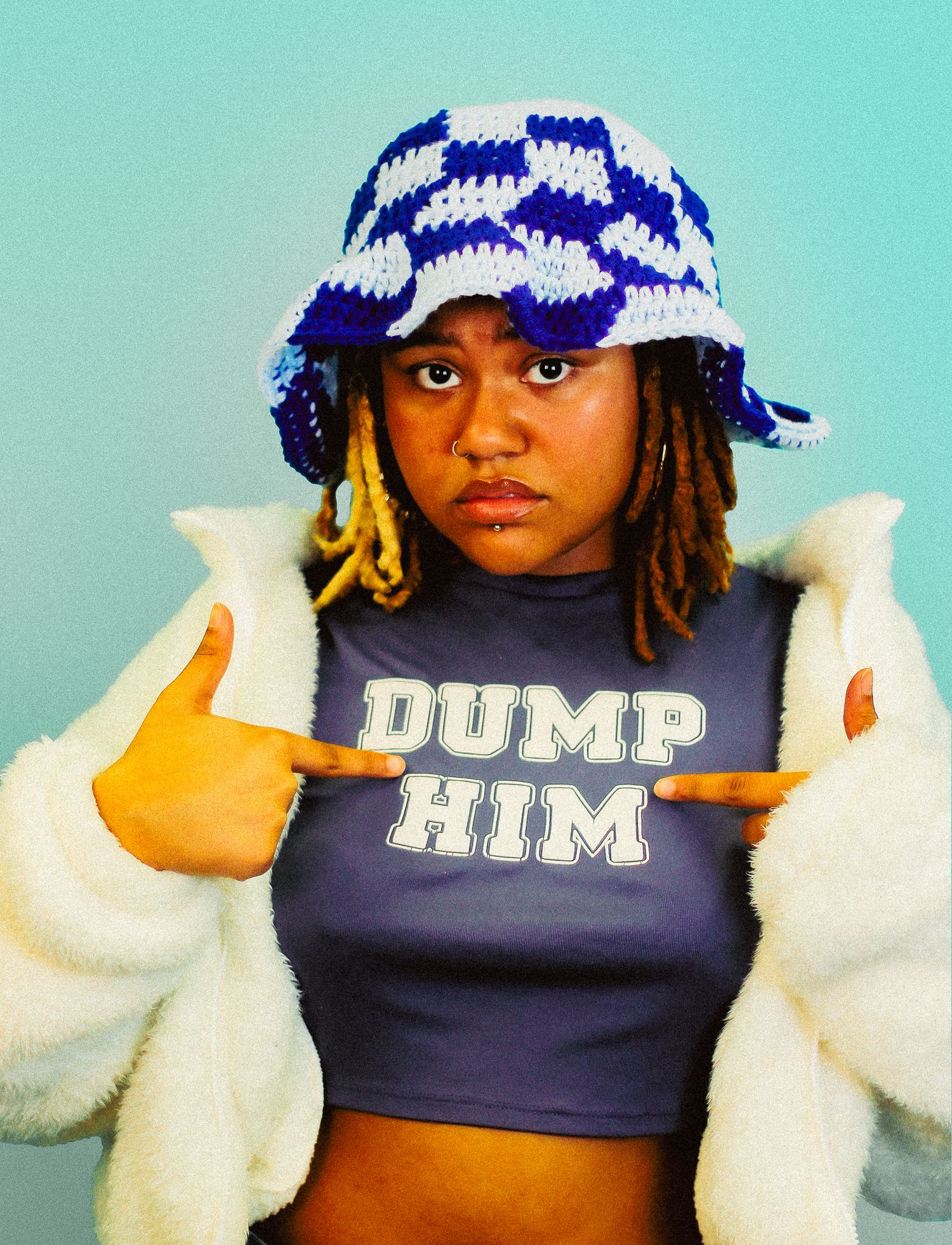 Young Black woman in blue checkered bucket hat points to her gray Dump Him crop top with white fluffy jacket.
