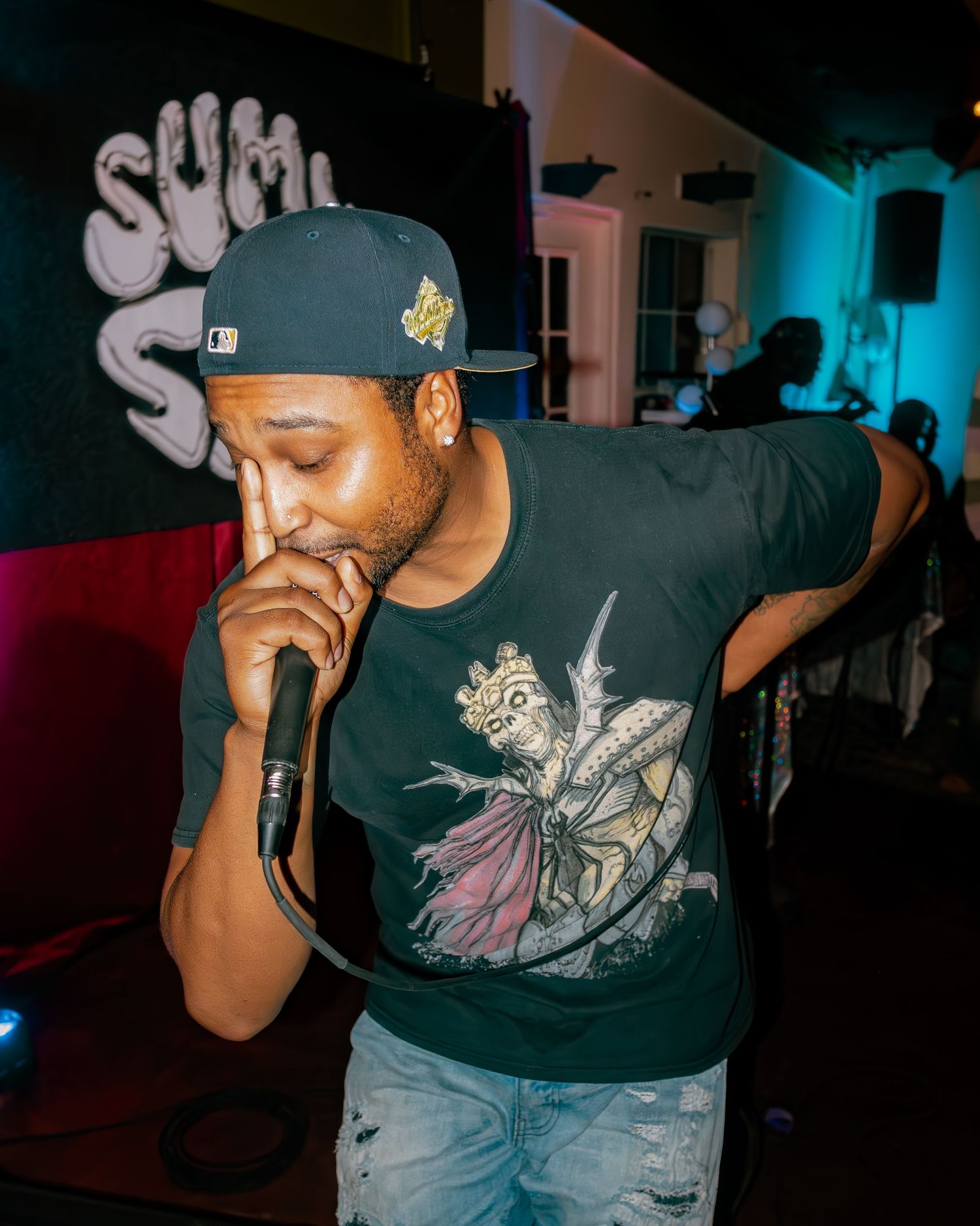 Close-up shot of performer wearing graphic t-shirt and baseball cap in dark venue with blue lighting.