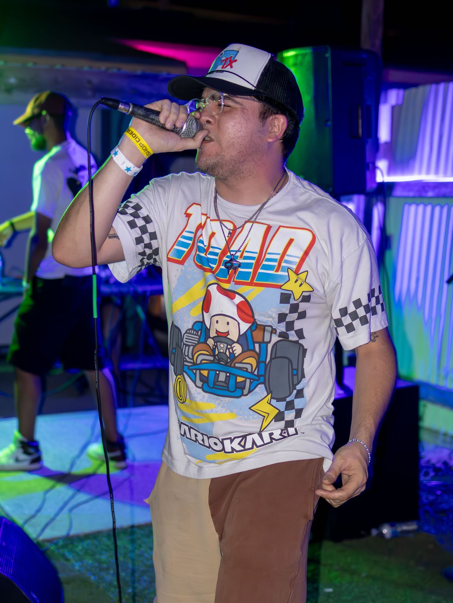 Performers in white t-shirts with checkered patterns sing on stage under colorful nightclub lighting.