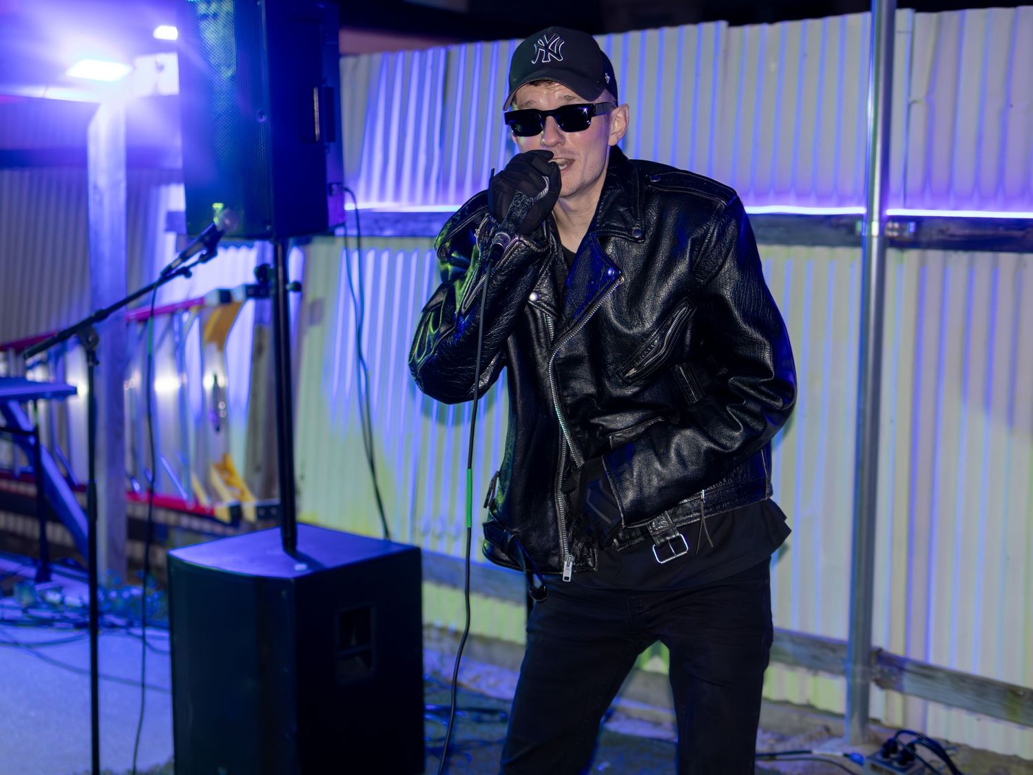 Concert performer in dark clothing and sunglasses poses dramatically under blue stage lights.