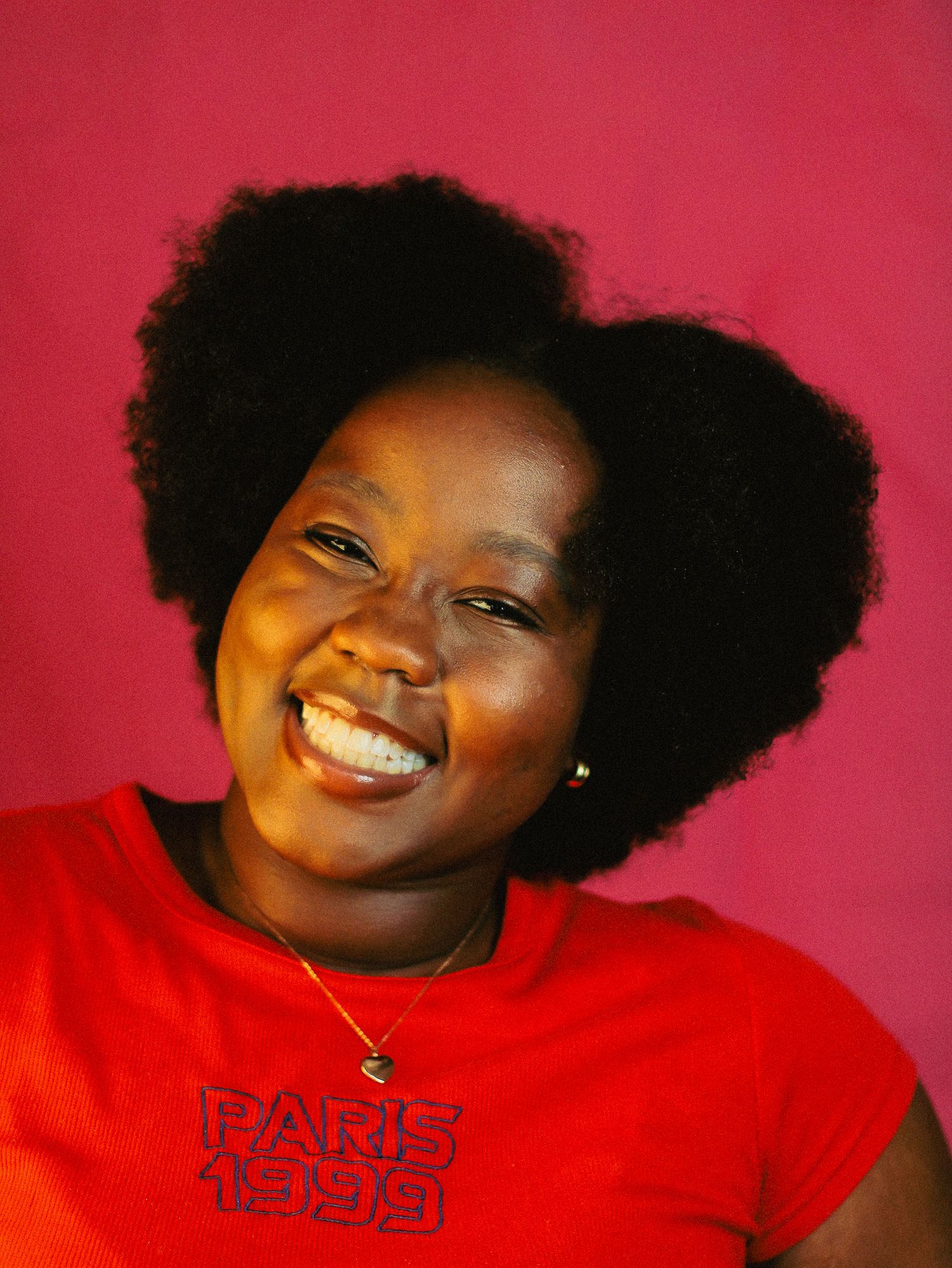 Joyful Black woman with natural afro hairstyle wearing a red Paris 1999 tee, laughing against pink background.