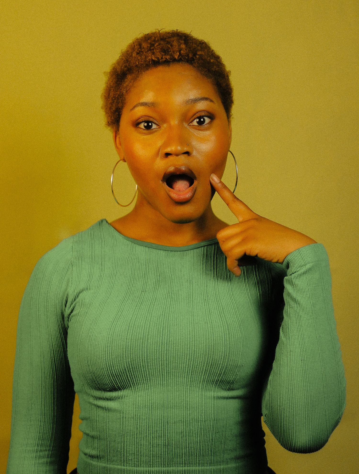 Young woman with short natural hair making surprised expression, pointing to cheek, wearing teal ribbed top.