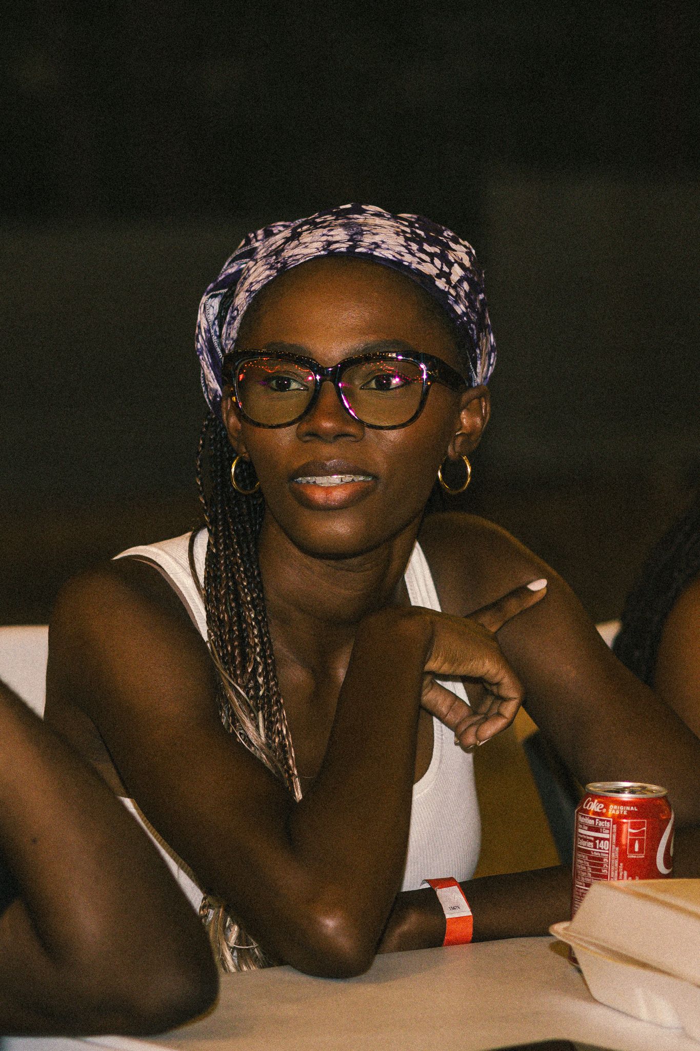 A person wearing glasses and braids smiles while sitting at a table with a Coca-Cola can.