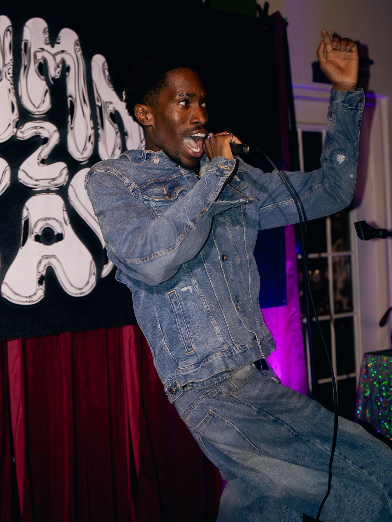 Performer in denim jacket dances energetically on stage with colorful lighting and curtains in the background.