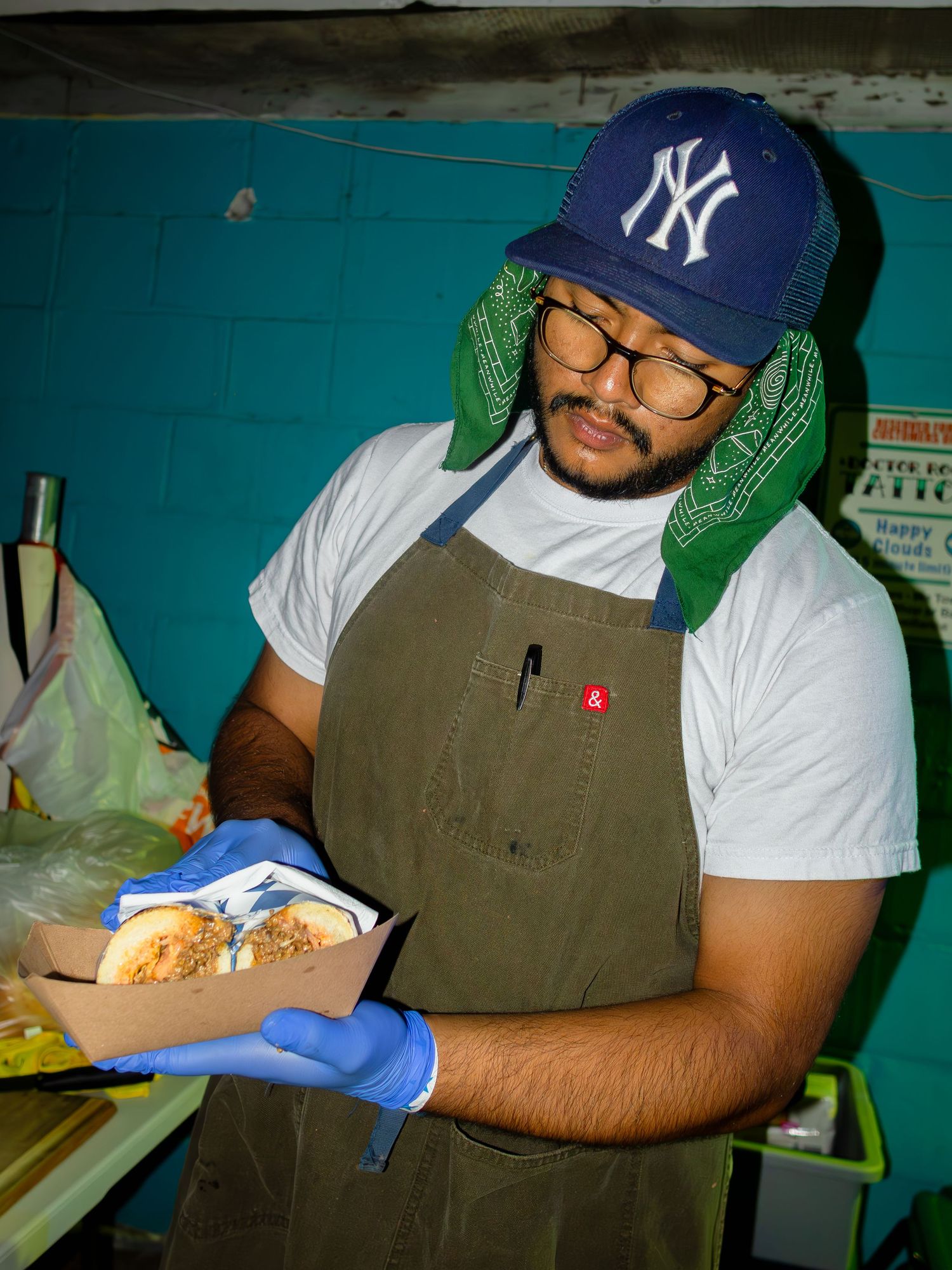 Person in Yankees cap and overalls handling food with blue gloves in commercial kitchen.