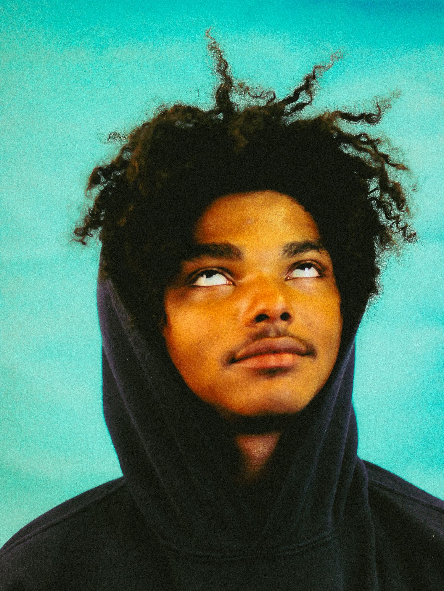 Young man with curly natural hair in dark hoodie looking upward against turquoise background.