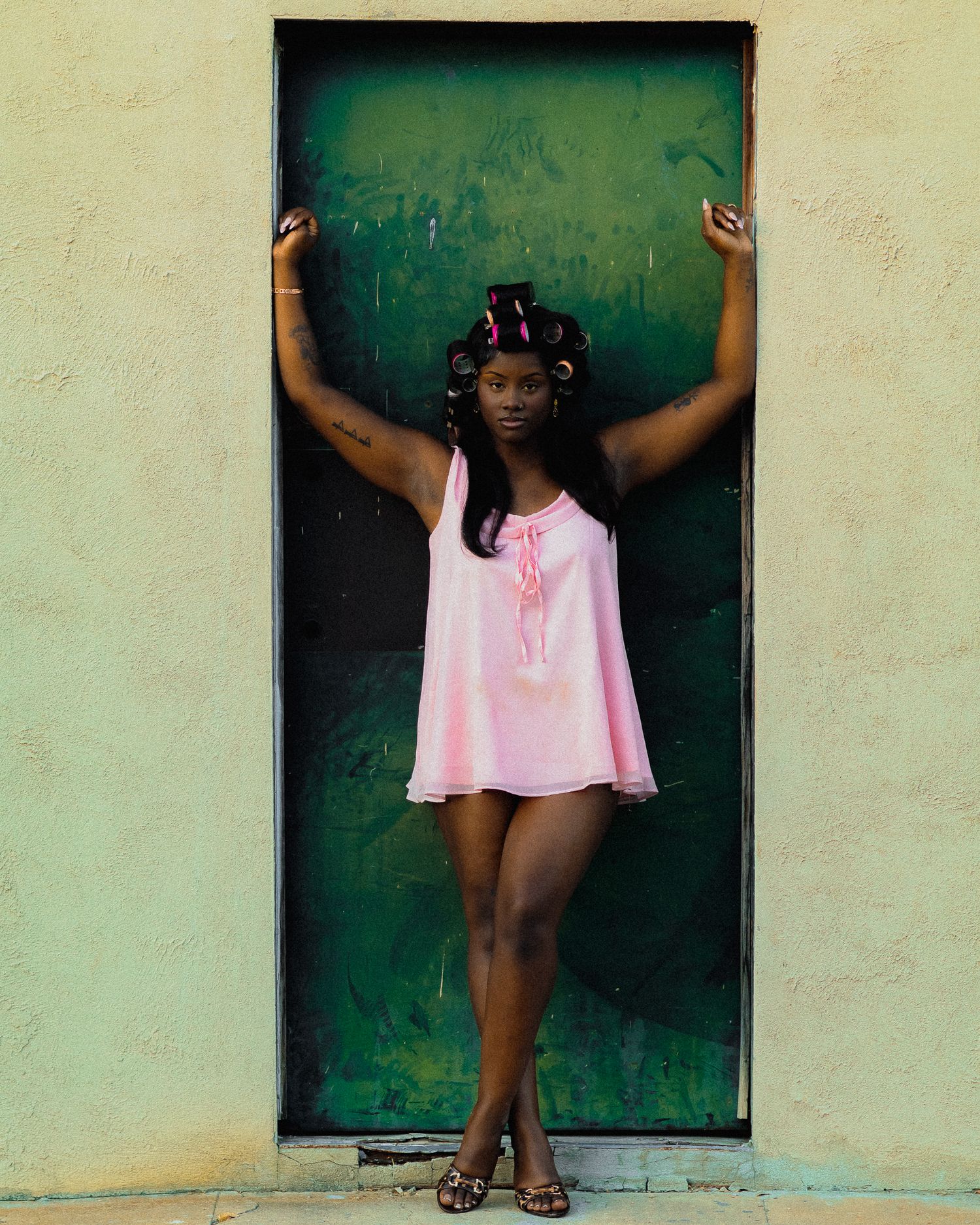 Model in pink dress poses playfully against green door.