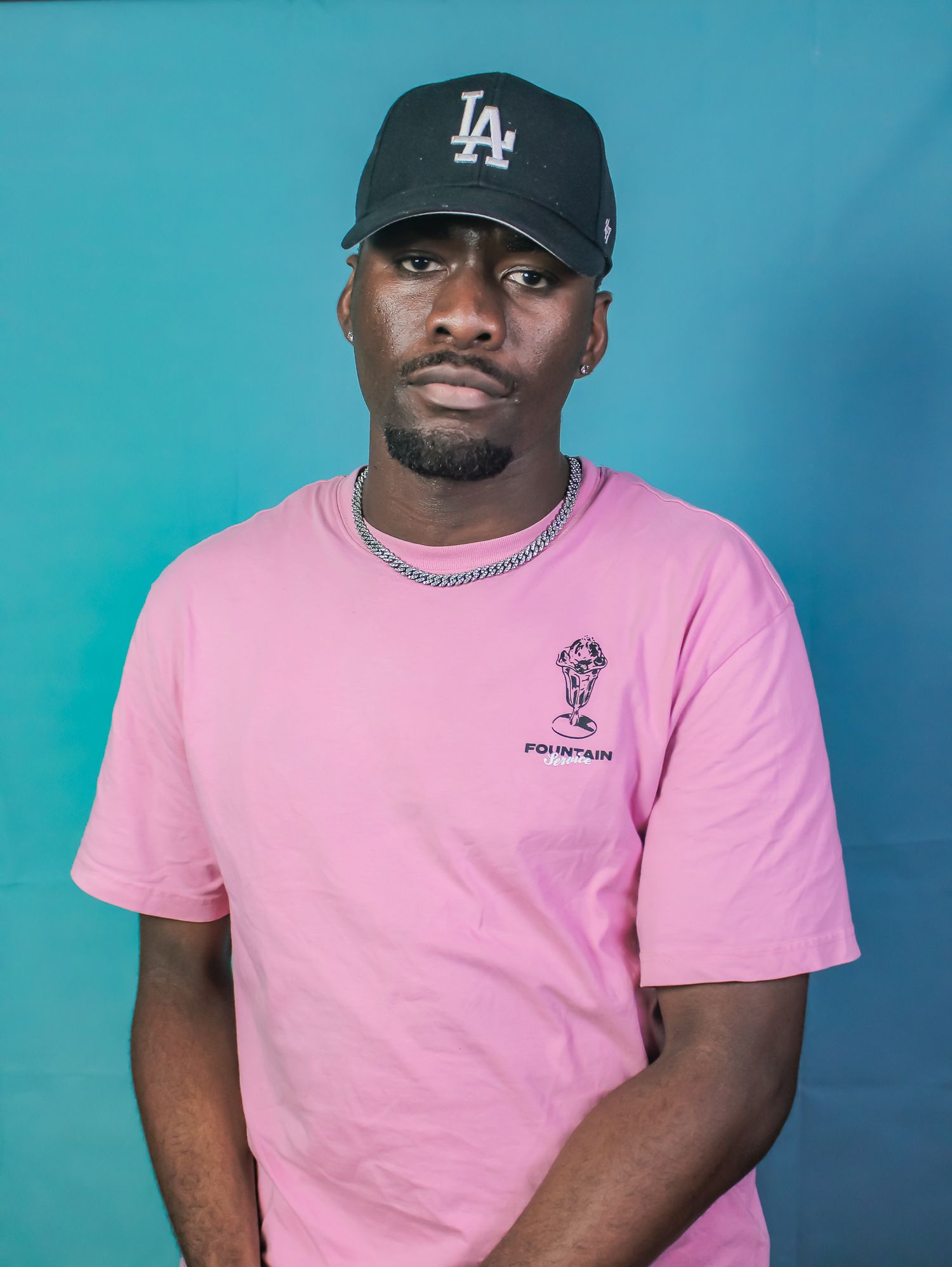 Young Black man in pink Fountain tee and LA Dodgers cap poses against teal background.