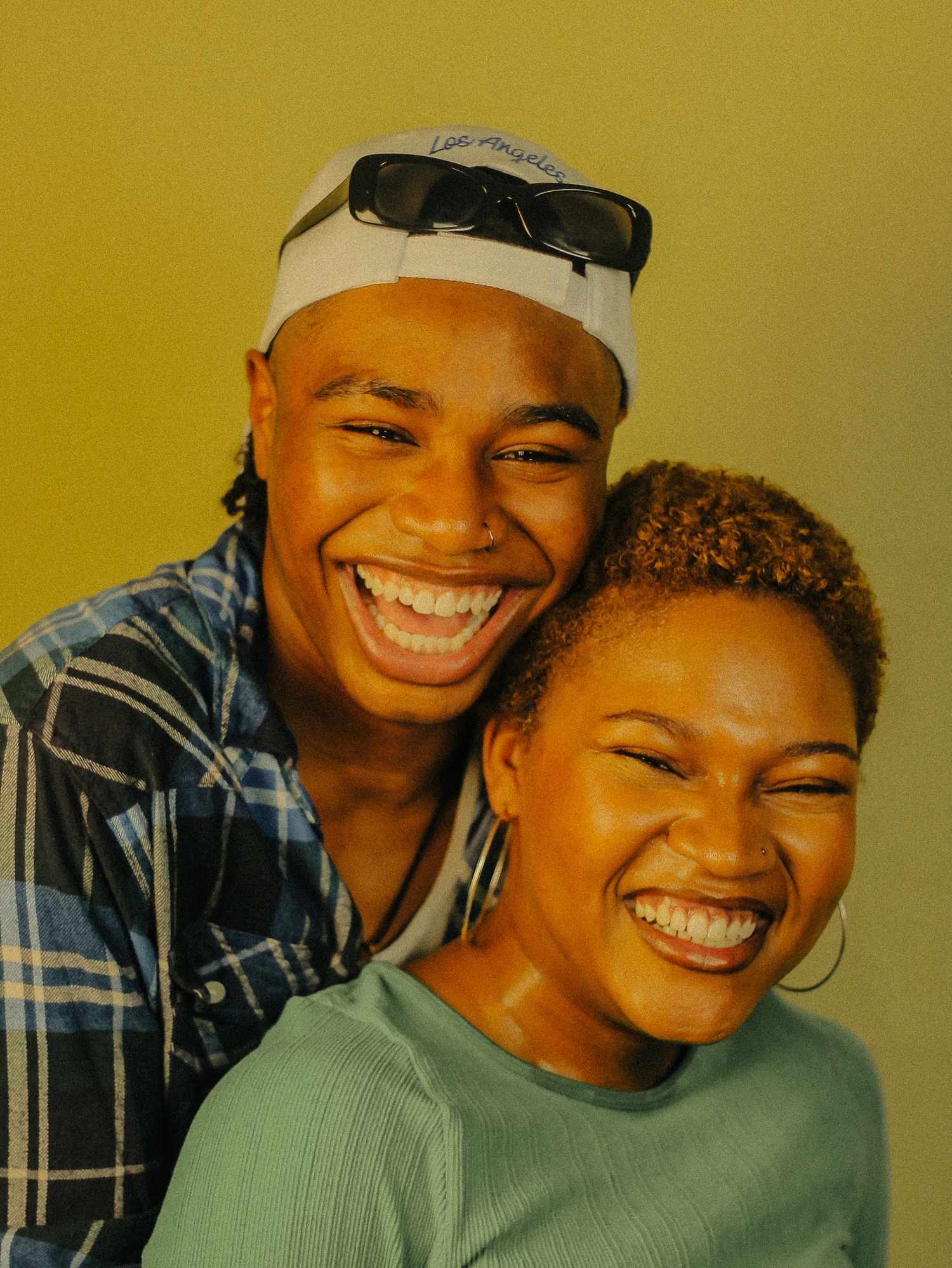 Two young people laughing joyfully together, one wearing a white LA cap and blue plaid shirt.