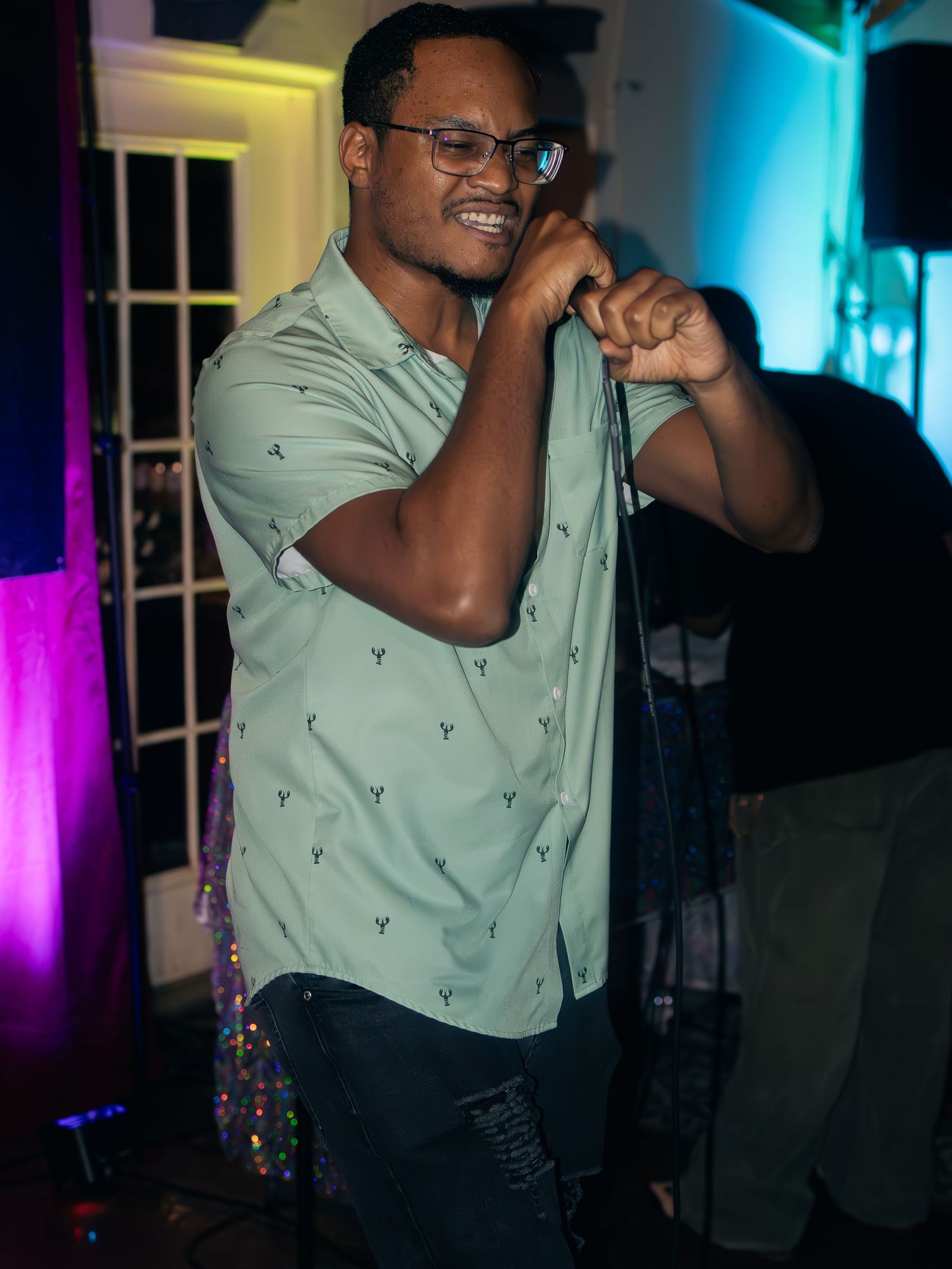 Artist in mint colored shirt performs expressively with glasses under dramatic stage lighting.