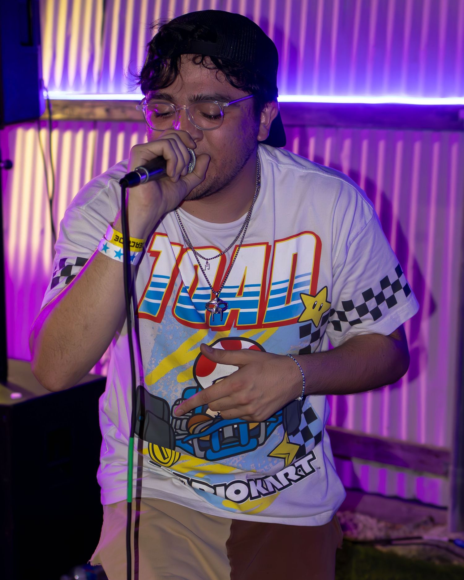 Performer in a white Mario Kart t-shirt with checkered pattern singing on stage under purple lighting.