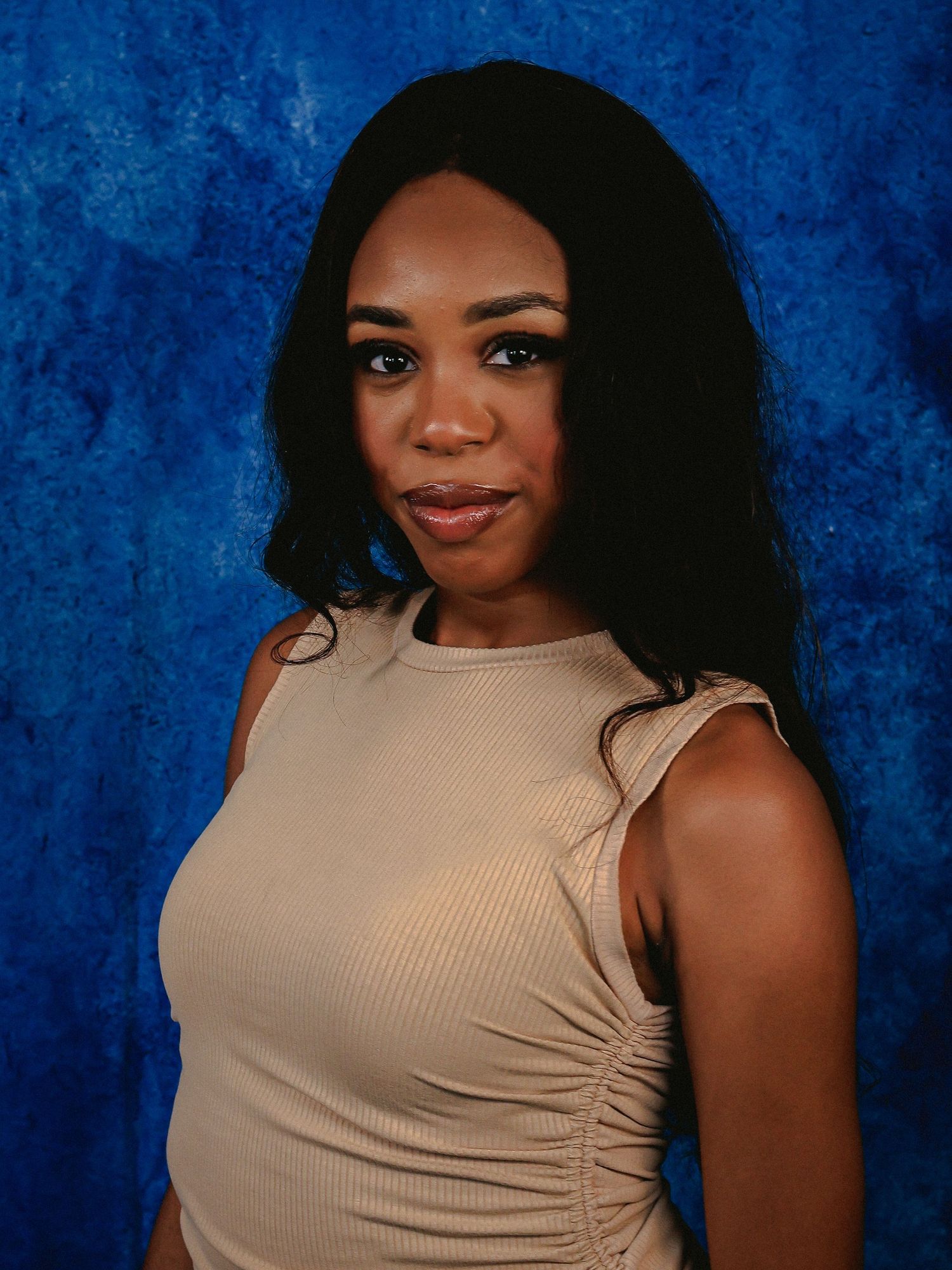 Woman in beige sleeveless top poses against textured blue background.