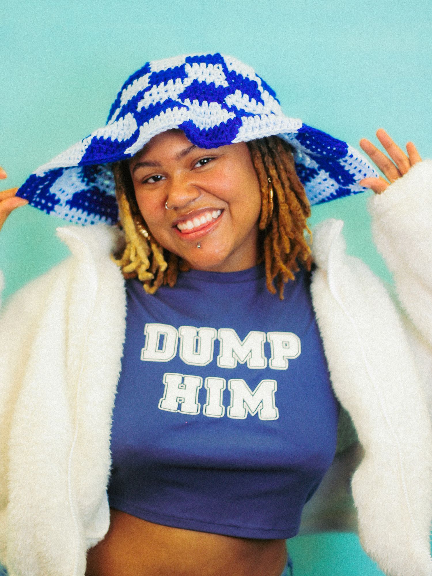 Smiling young woman wearing blue checkered crochet bucket hat and Dump Him graphic crop top poses playfully.