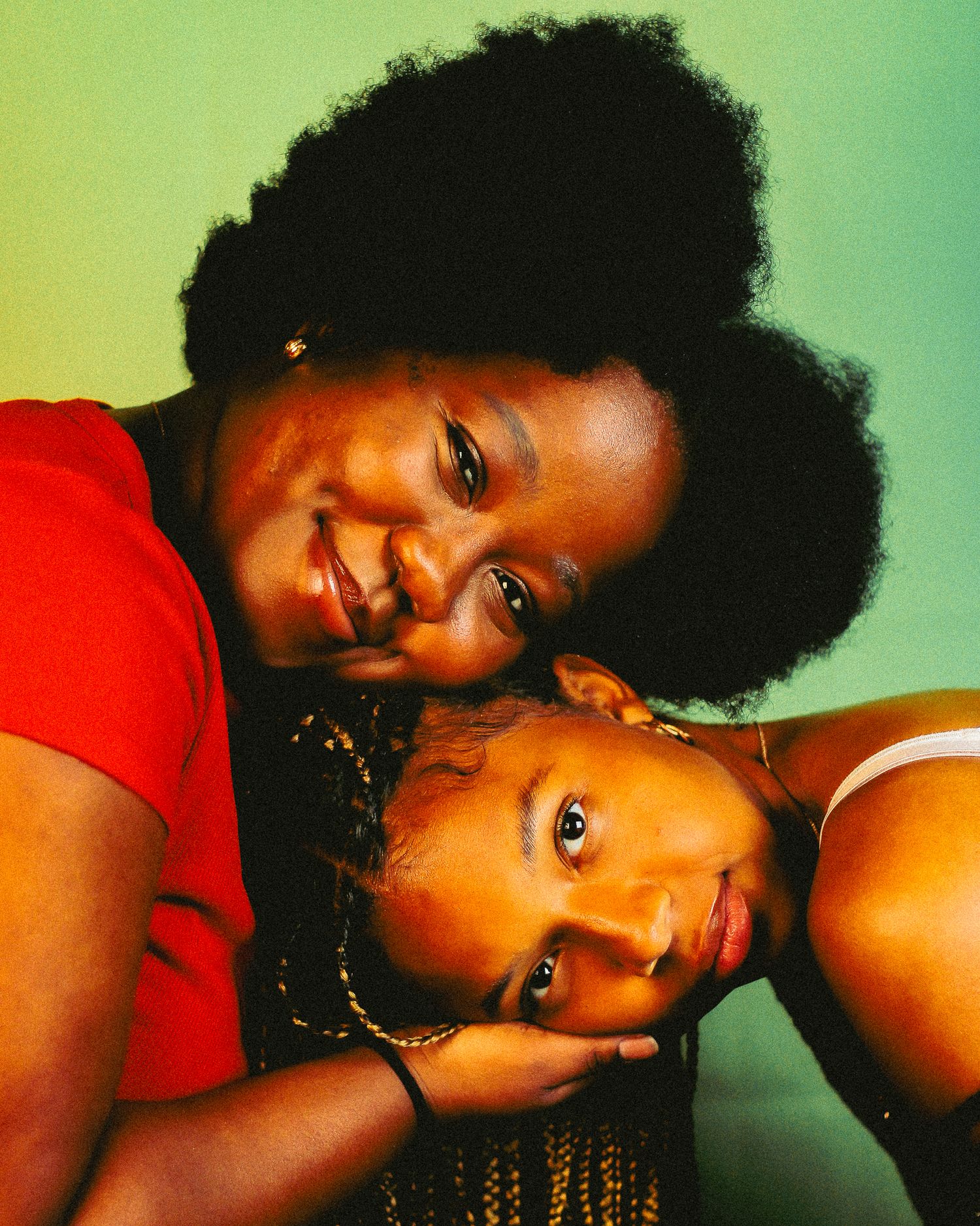 Two Black women smiling warmly in close portrait, one with natural afro in red top leaning over other.