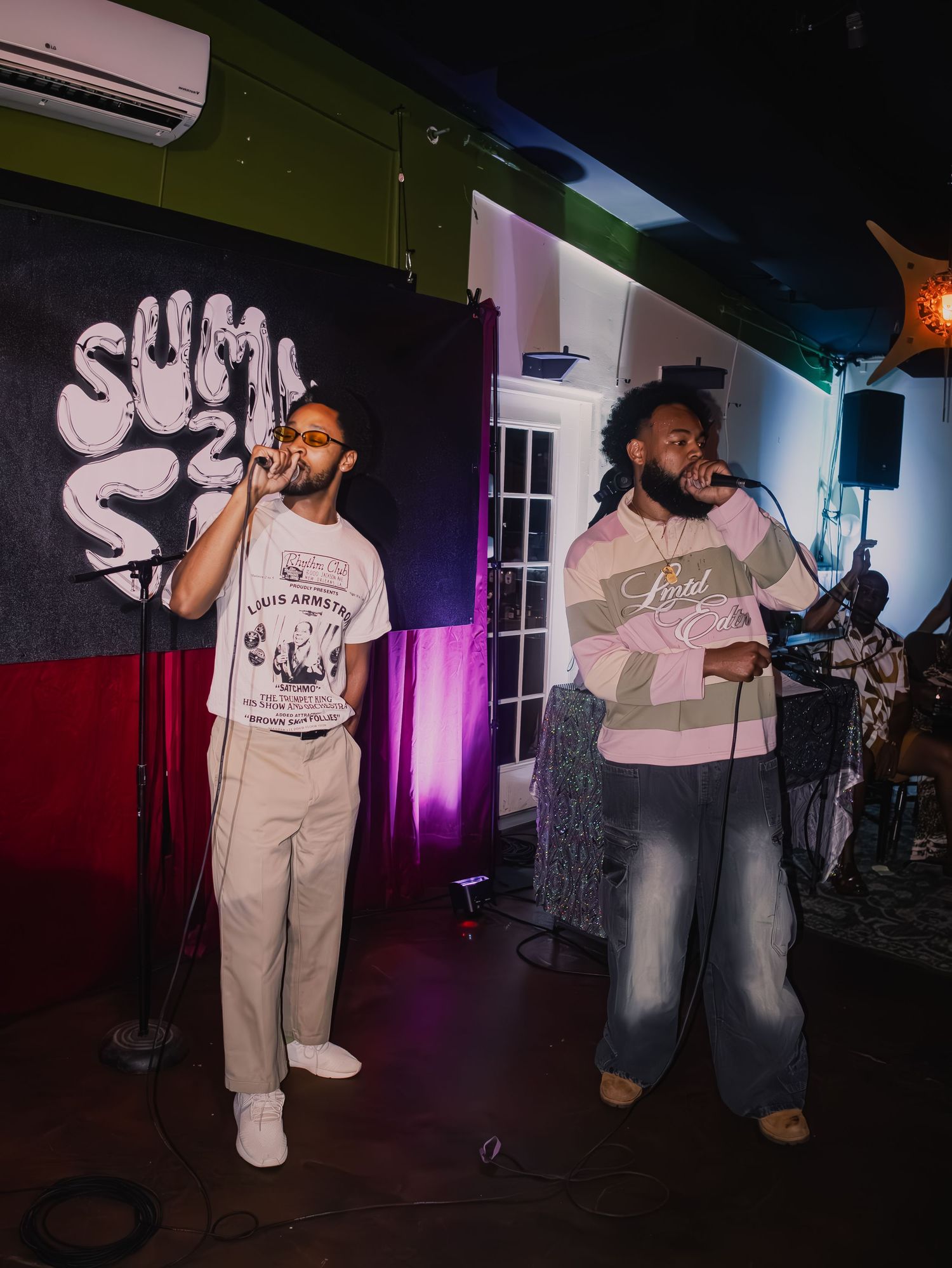Two performers sharing stage with black and red banner backdrop during live show.