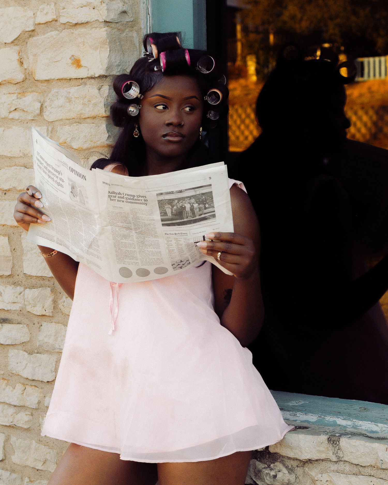 A person wearing hair rollers poses with a newspaper against a stone wall background.