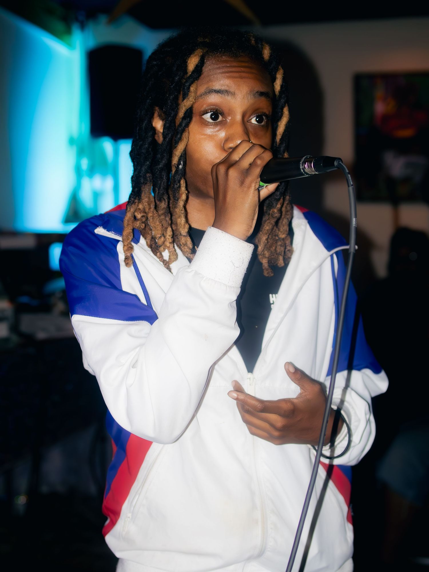 Artist performing with microphone wearing white jacket with blue accents against dark background.
